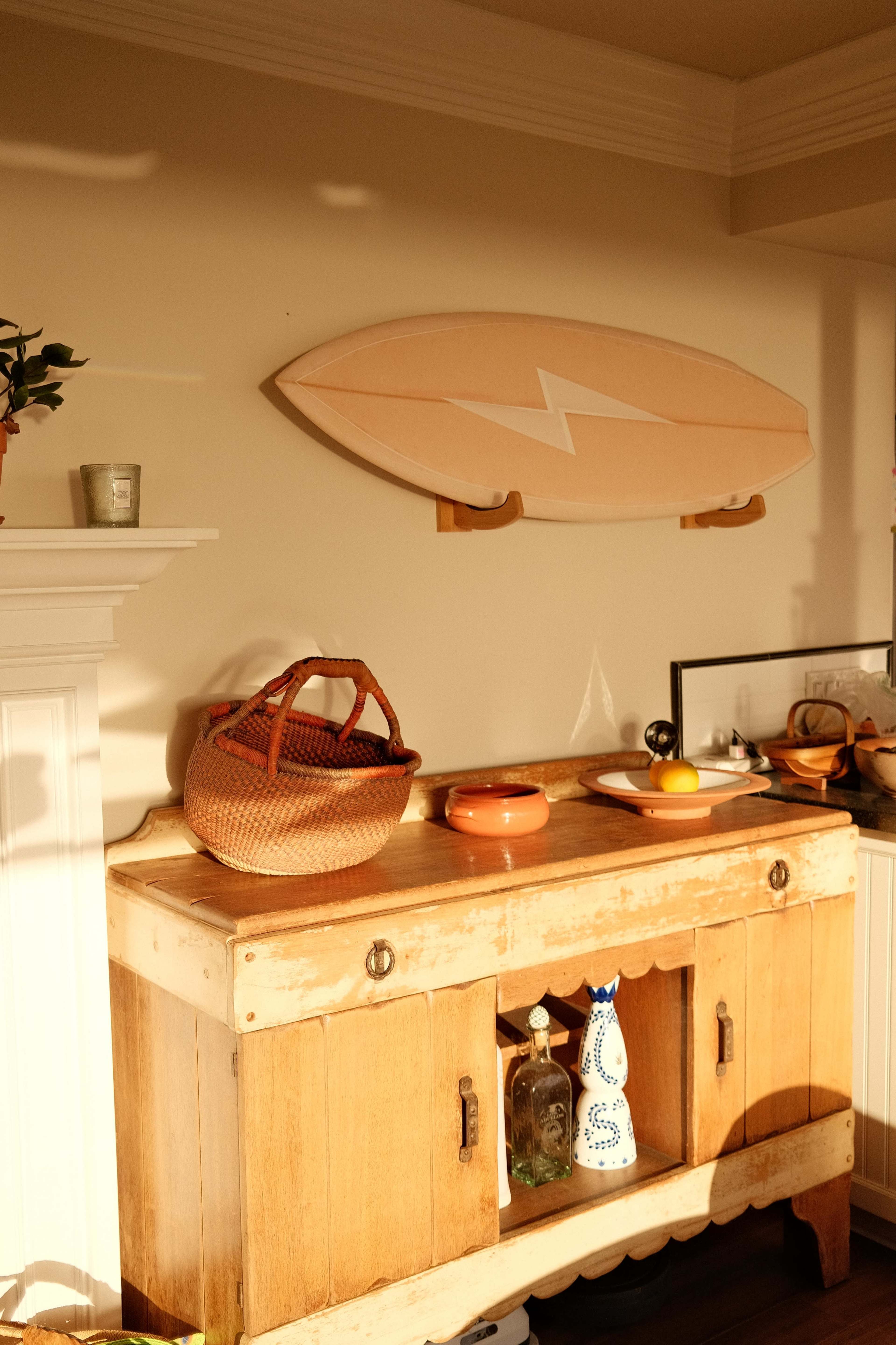 A wooden sideboard with a glass display and a woven basket sits beneath a surfboard mounted on the wall.