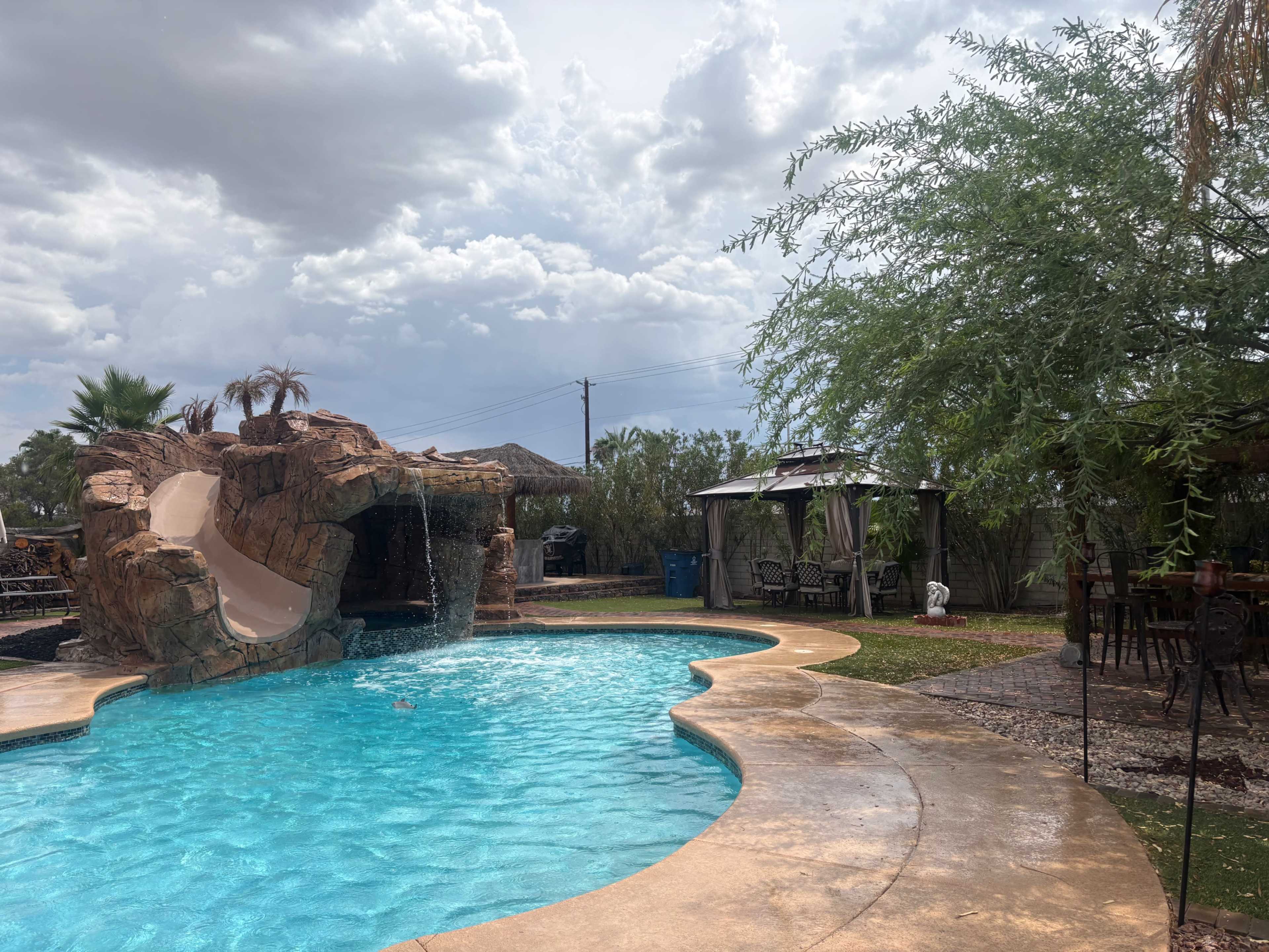 The image shows a swimming pool with a rock formation and a waterslide, surrounded by palm trees and outdoor seating areas under a cloudy sky.