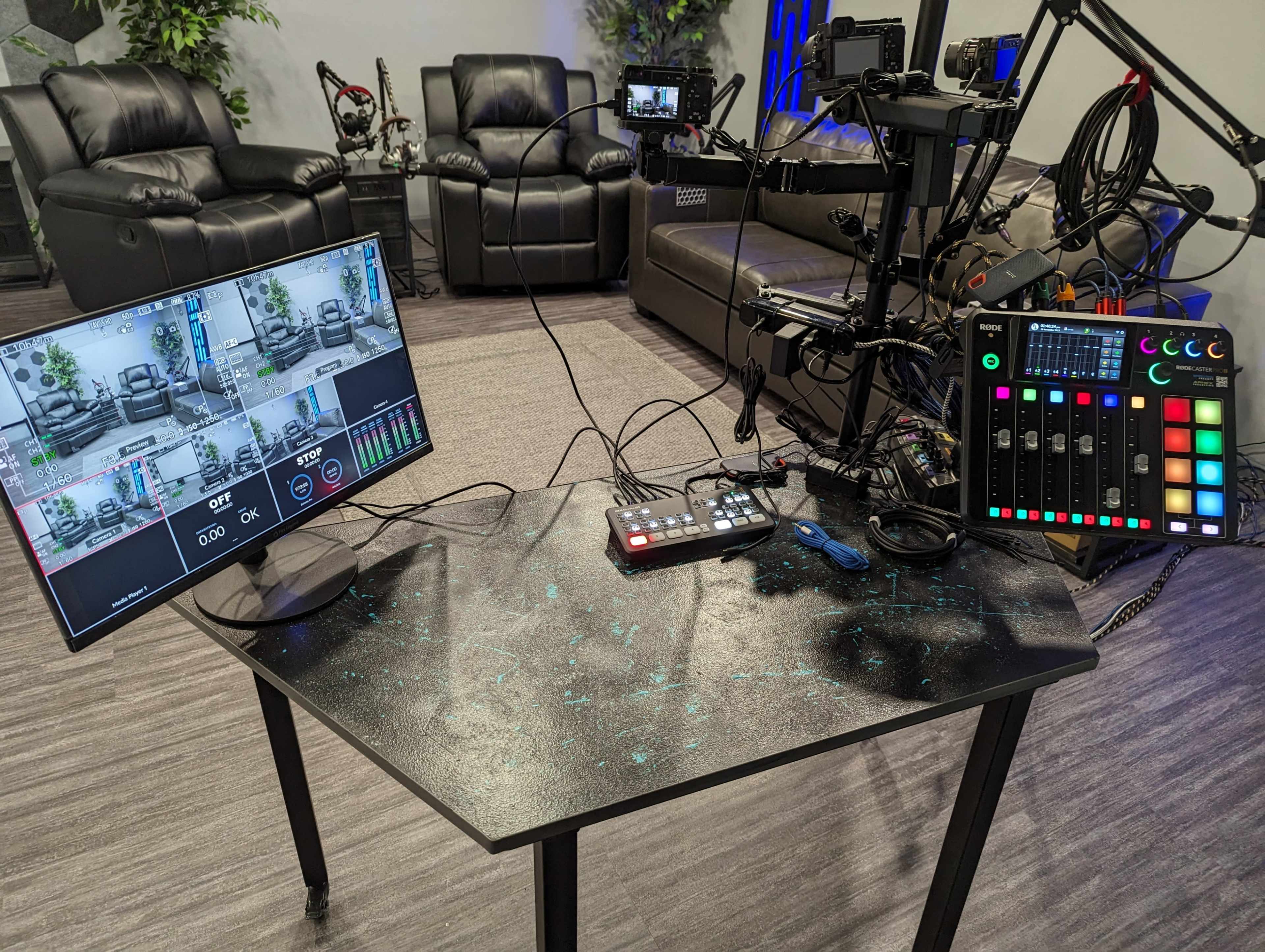 A control setup featuring a monitor displaying multiple camera feeds, a video switcher, and a messy arrangement of cables on a table in a studio environment.