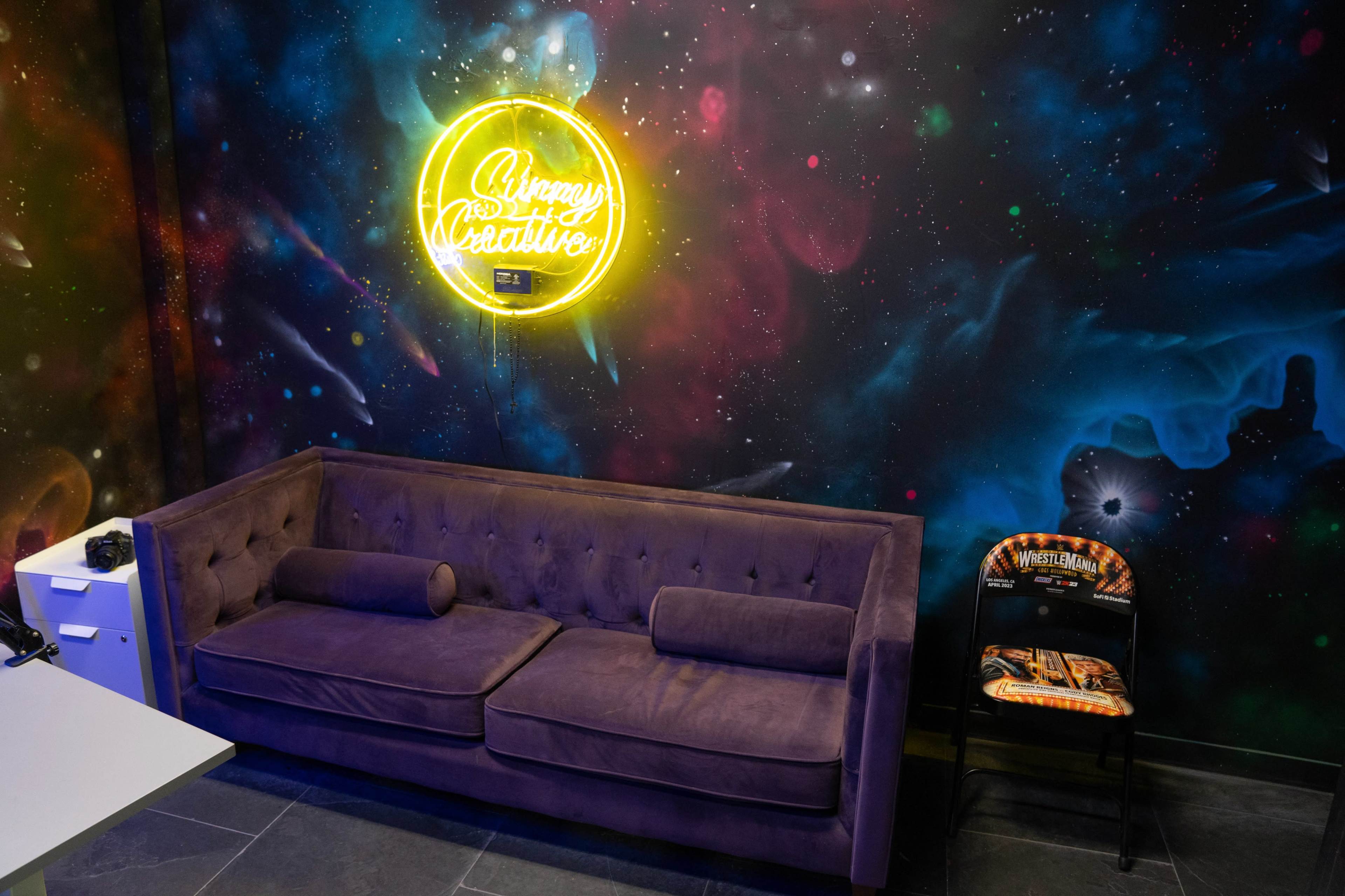 A purple couch against a vibrant, galaxy-themed wall with a neon sign that reads "Always Be Creative."