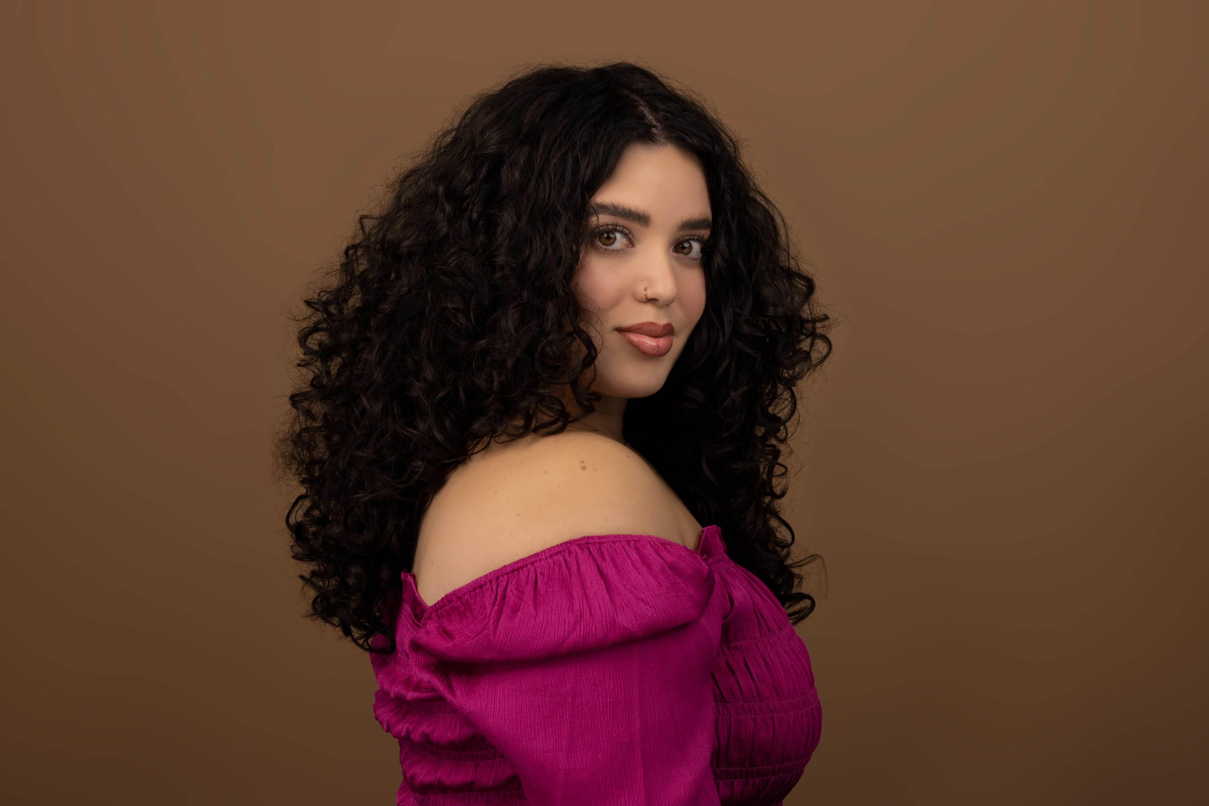 A woman with curly black hair poses in profile against a brown background while wearing an off-the-shoulder pink top.