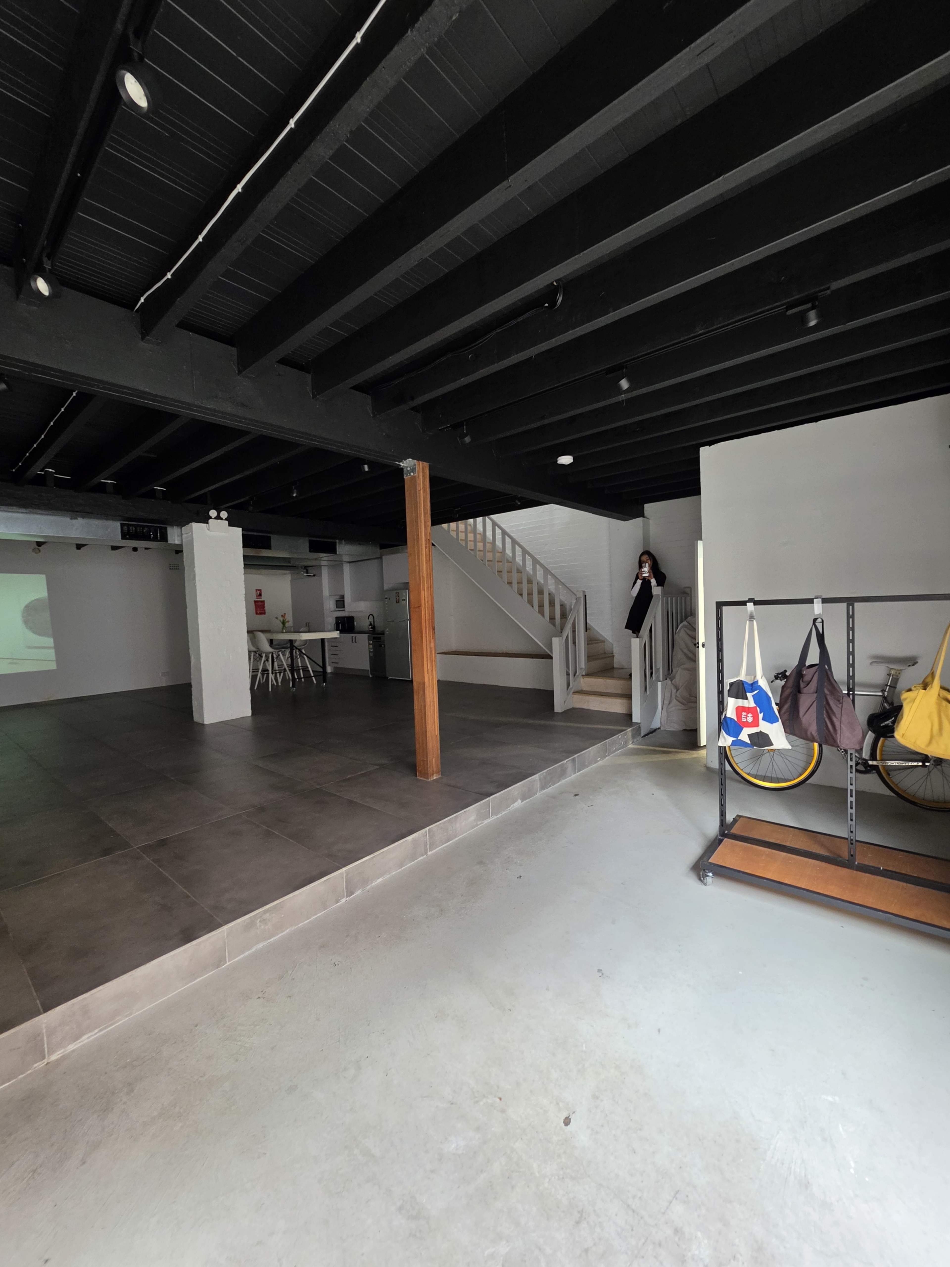 The image shows a spacious interior of a building with a tiled floor, wooden beams, a staircase, and bicycles on a rack.