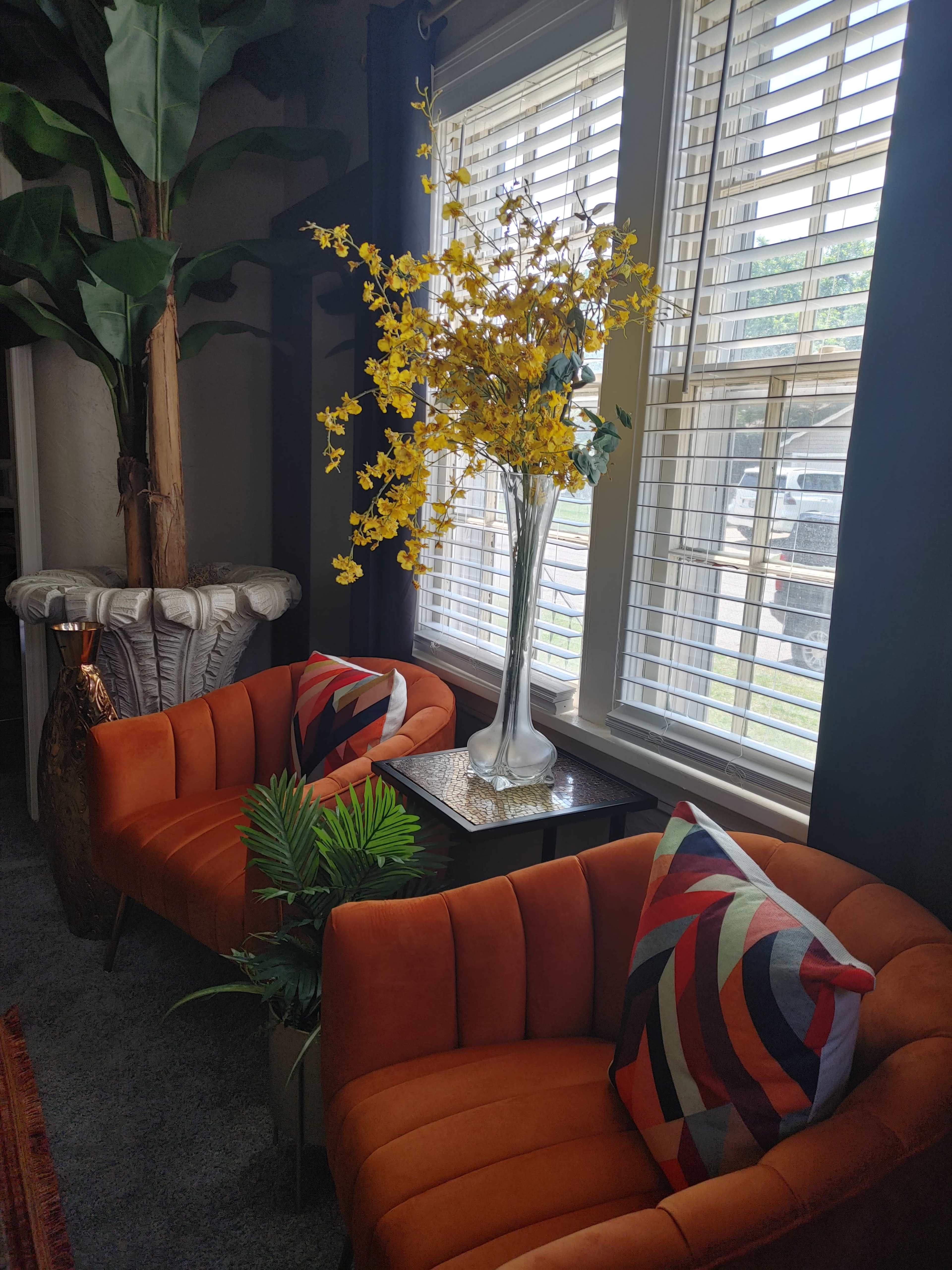 The image shows two orange chairs with geometric pillows beside a glass table, a large vase of yellow flowers, and a tall plant in a decorative pot near a window with blinds.