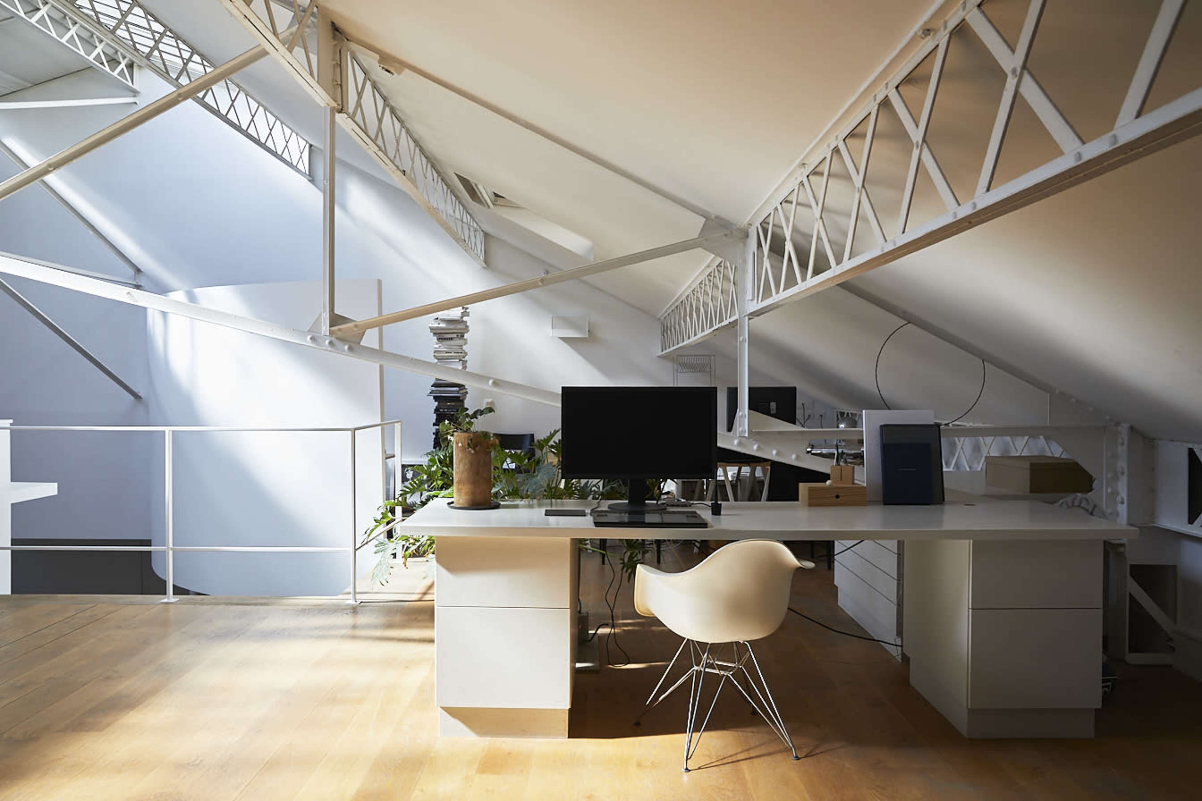 The image shows a modern office space with a desk, monitor, and an ergonomic chair, set under a sloped ceiling supported by metal beams and filled with natural light.