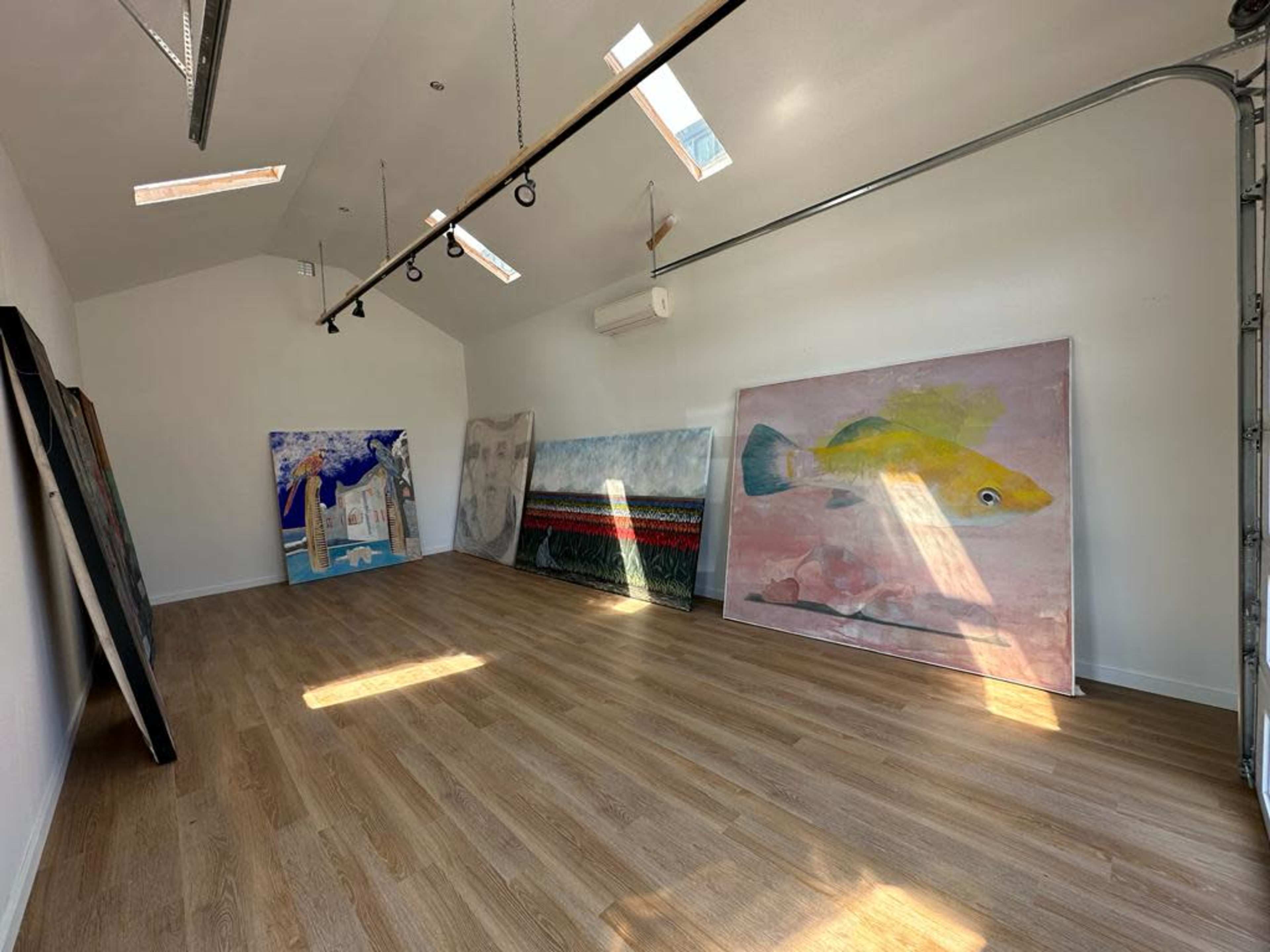 A spacious, well-lit room with wooden flooring, featuring several large paintings leaning against the walls.