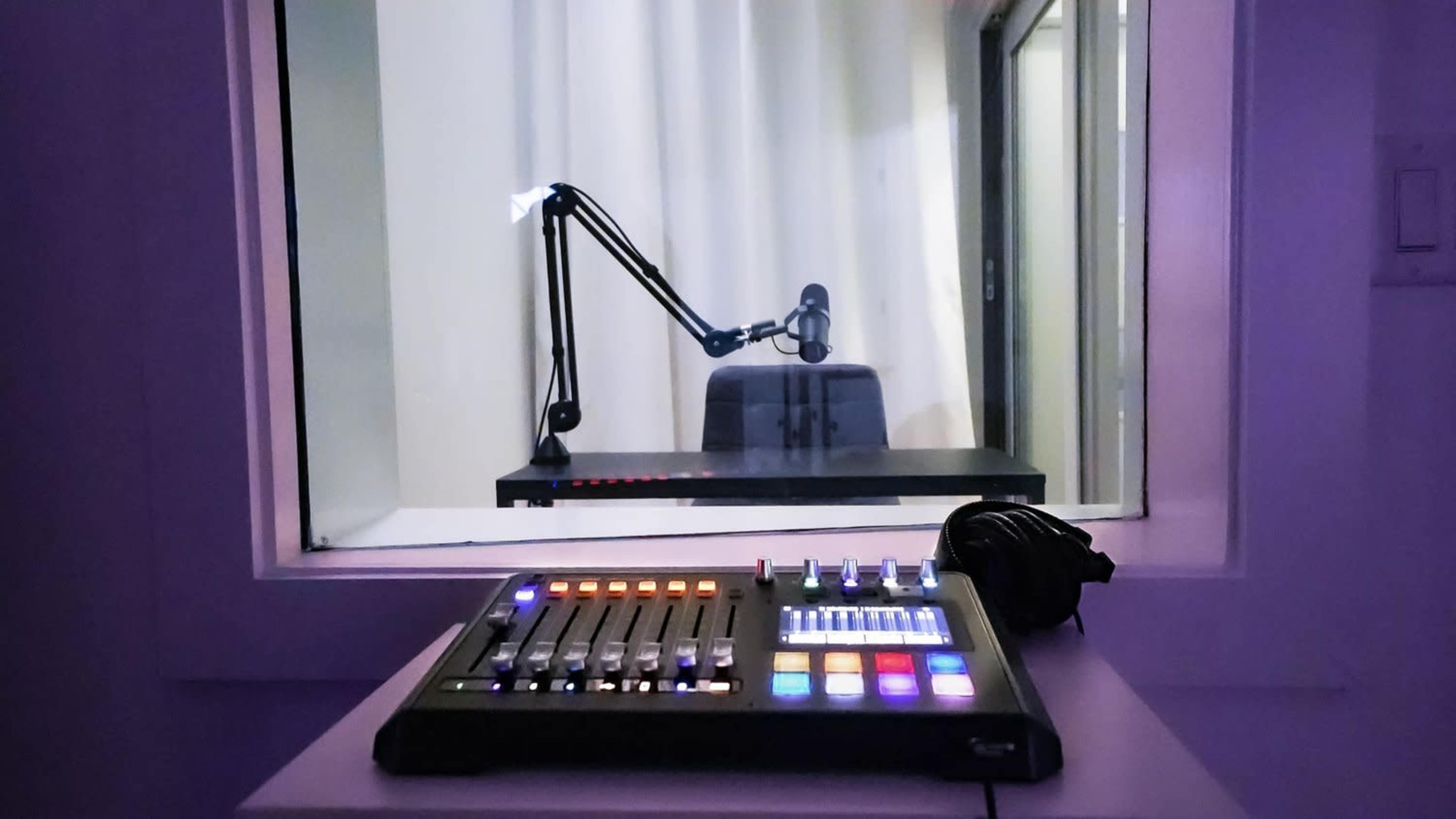 A control panel with various buttons and lights sits on a desk in the foreground, while a microphone arm and empty chair are visible through a glass window in the background.