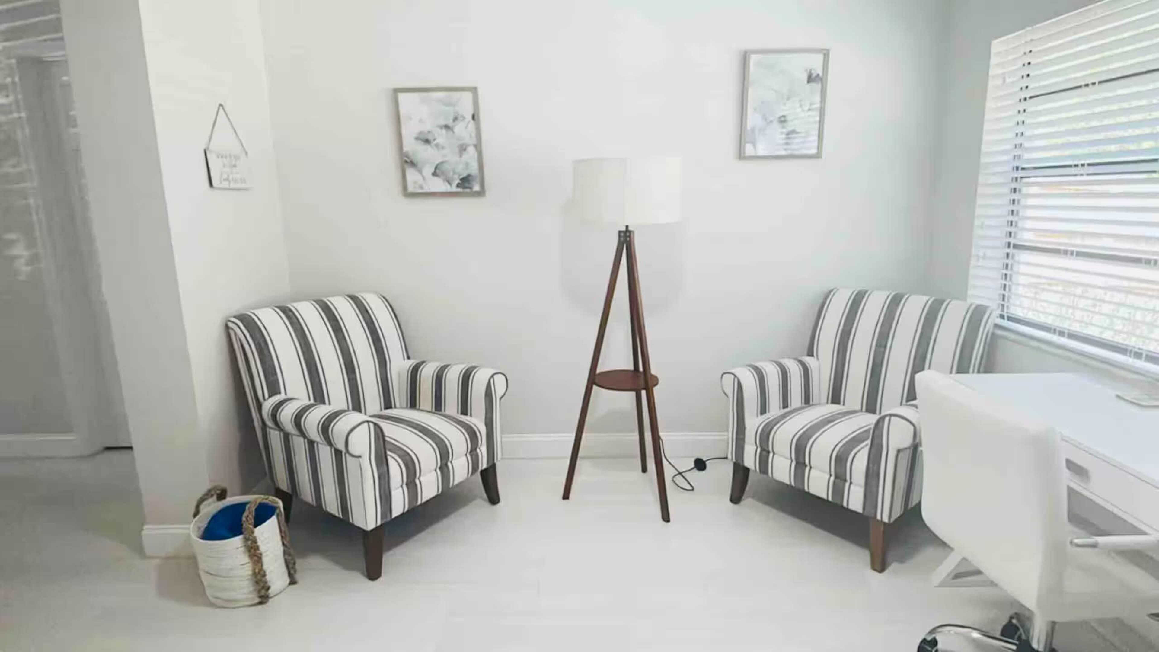 The image shows a minimalist room with two striped armchairs facing each other, a standing lamp, and two framed pictures on the wall.