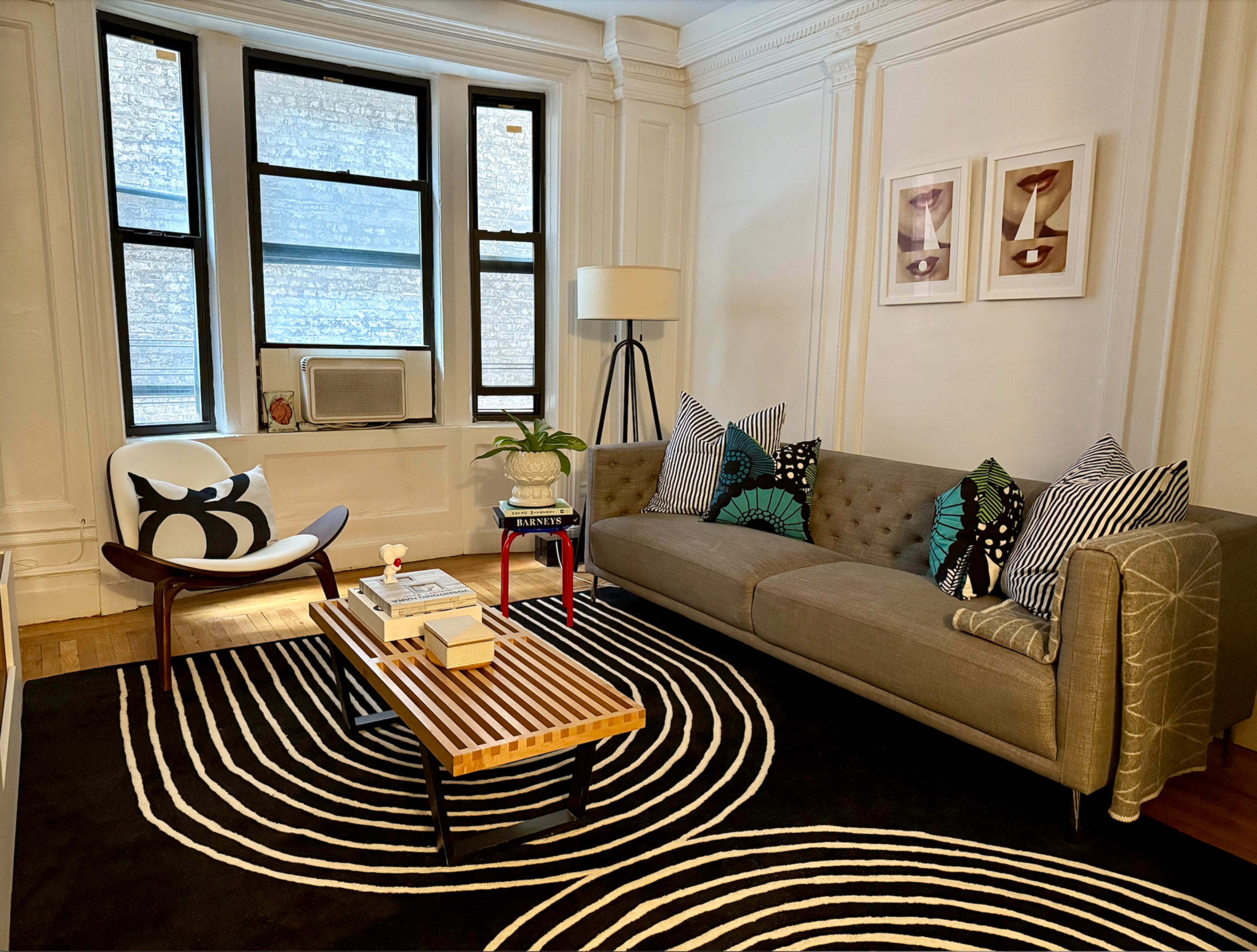 The living room features a gray tufted sofa, a striped area rug, a round coffee table, and large windows that provide natural light.