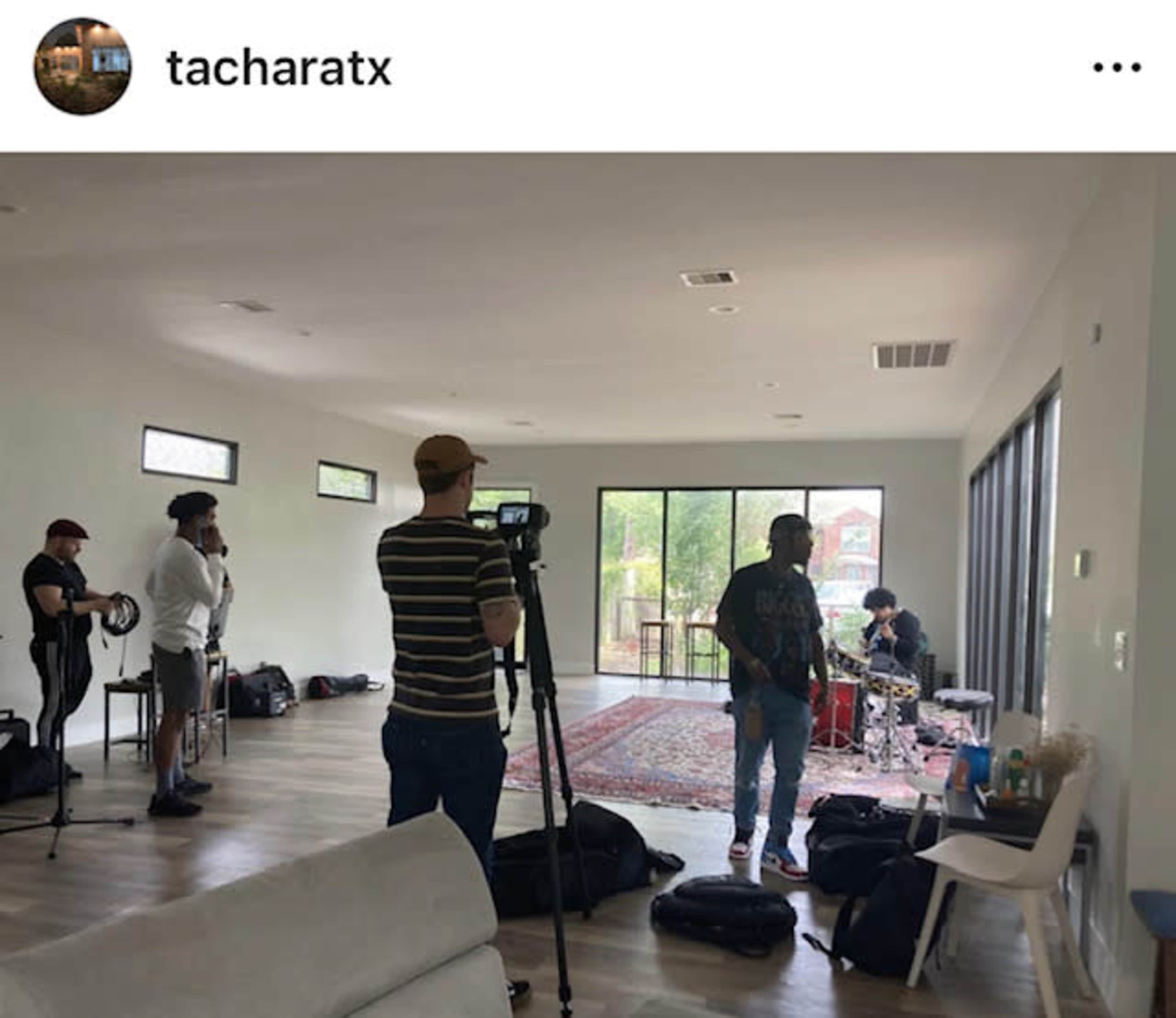 A group of people is filming a musician playing instruments in a spacious, modern room with large windows and minimal furniture.