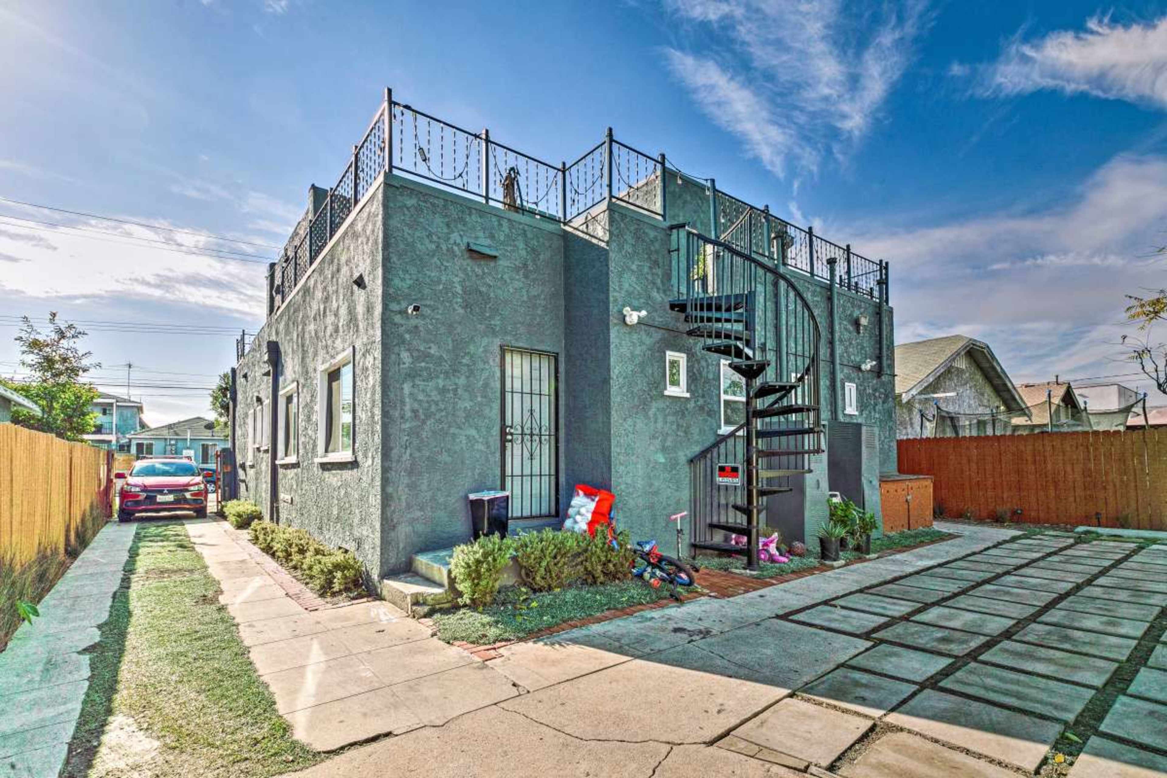 The image features a two-story gray building with a spiral staircase leading to a rooftop patio, surrounded by a green lawn and a gravel path.