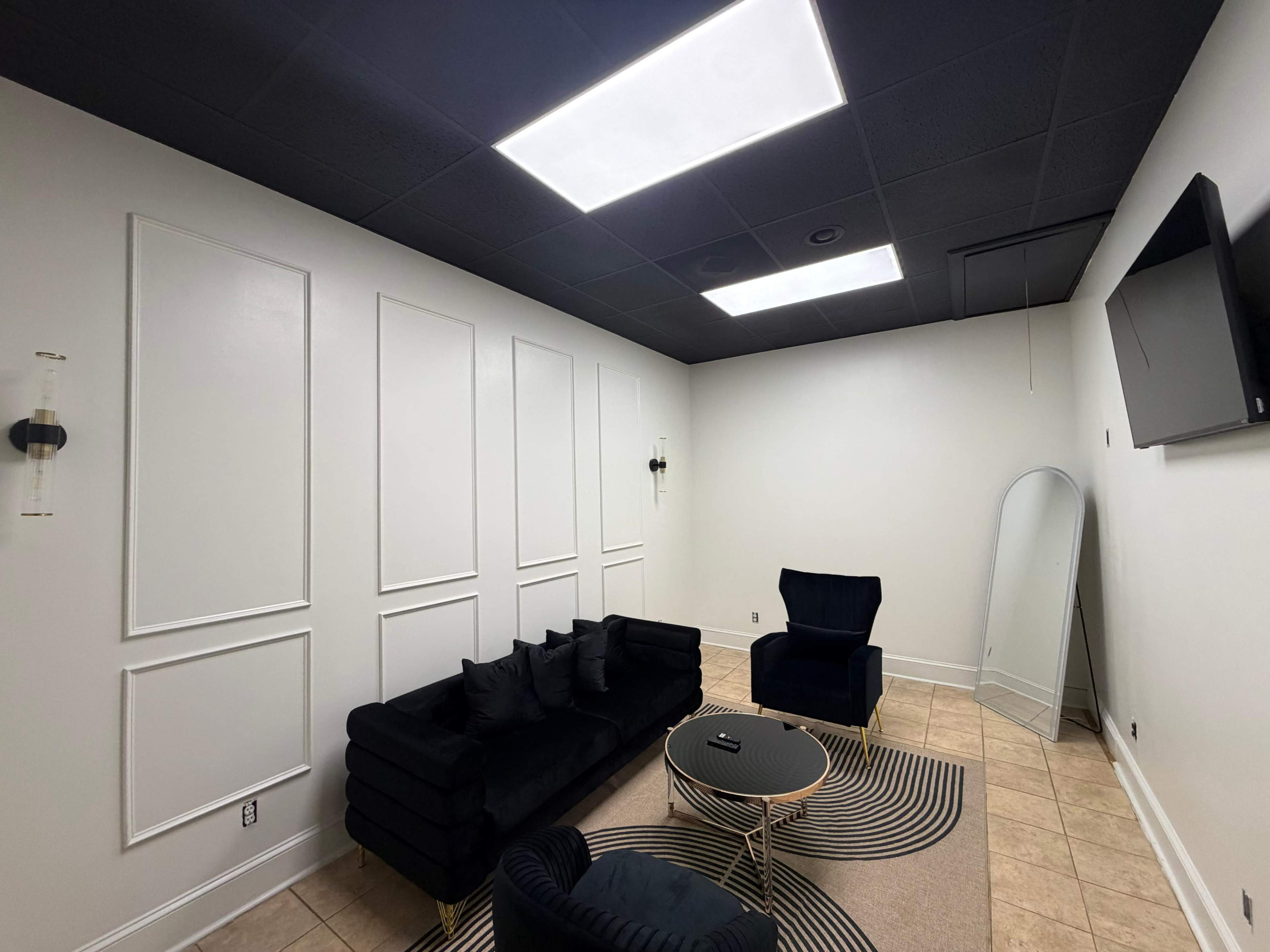 The image shows a modern, sparsely decorated room featuring a black sectional sofa, a blue armchair, a round area rug, a tall mirror, and a wall-mounted television.