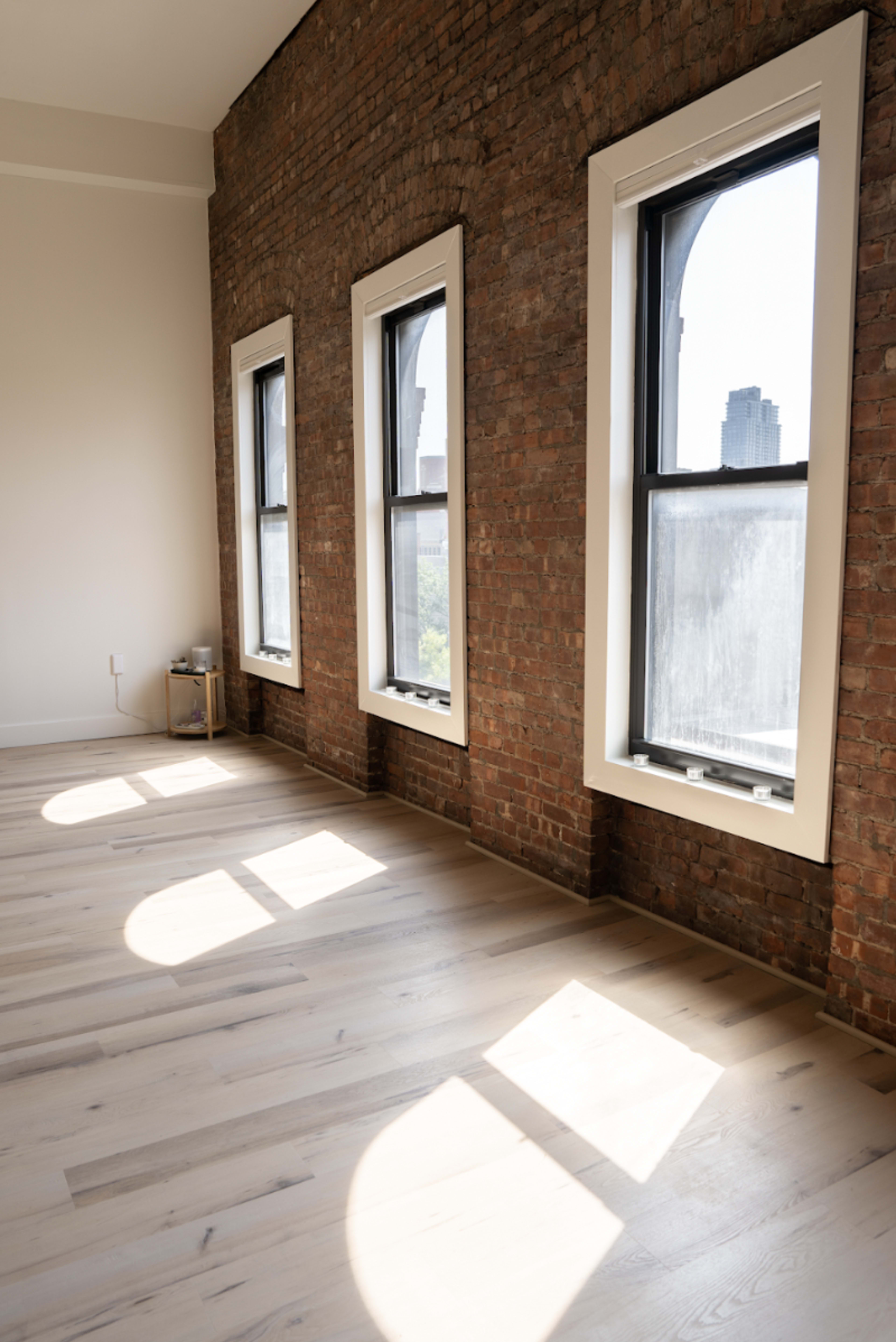 The image shows a room with exposed brick walls, large windows, and sunlight casting shadows onto the light-colored wooden floor.