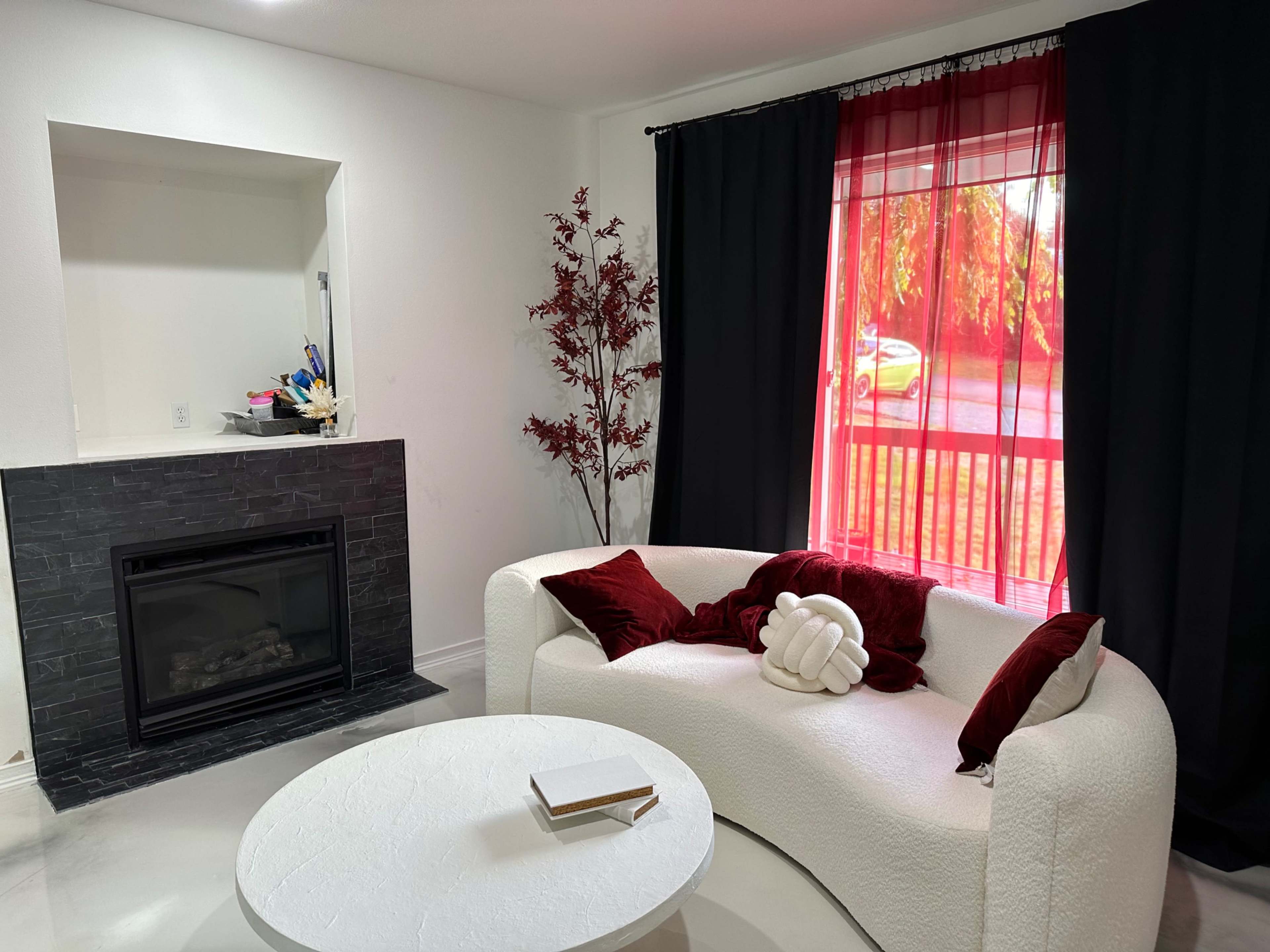 A white modern couch with red and white cushions, positioned near a circular coffee table in a room featuring a fireplace and large windows with black curtains.