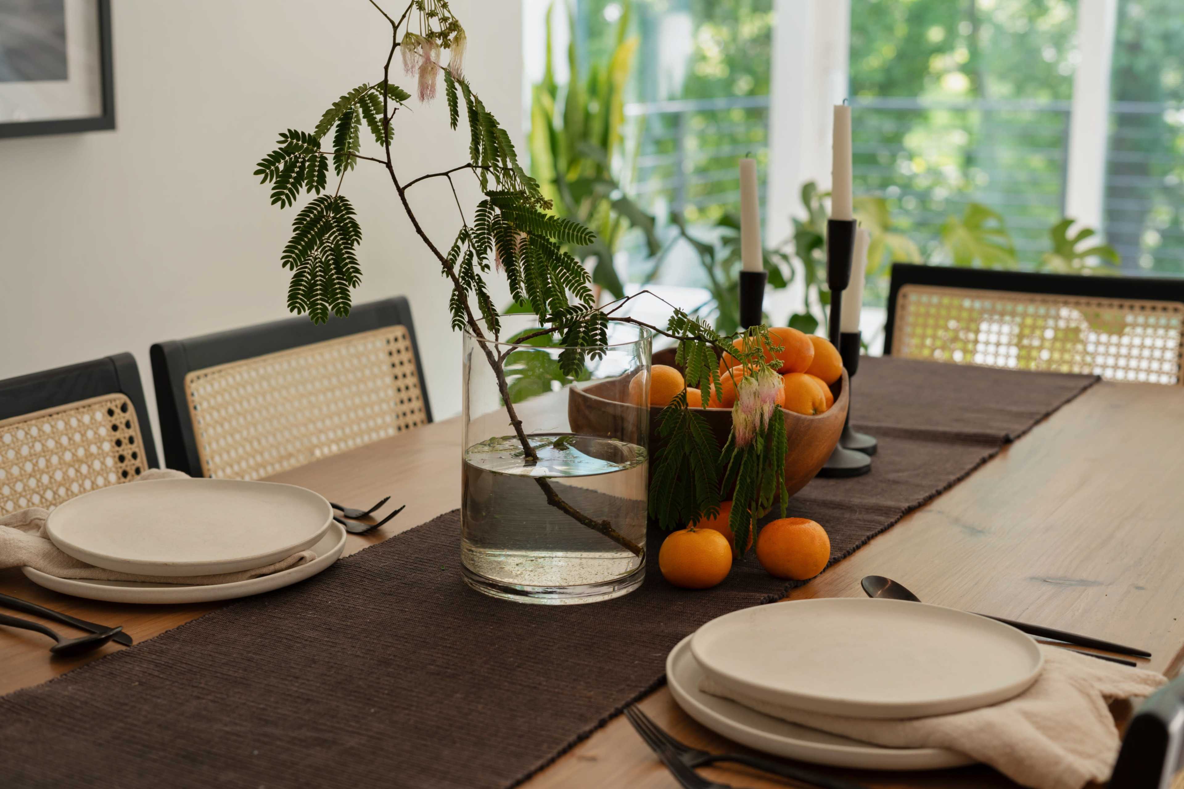 A dining table is set with natural decorations, including a glass vase with a plant, bowls of oranges, and simple dishware.