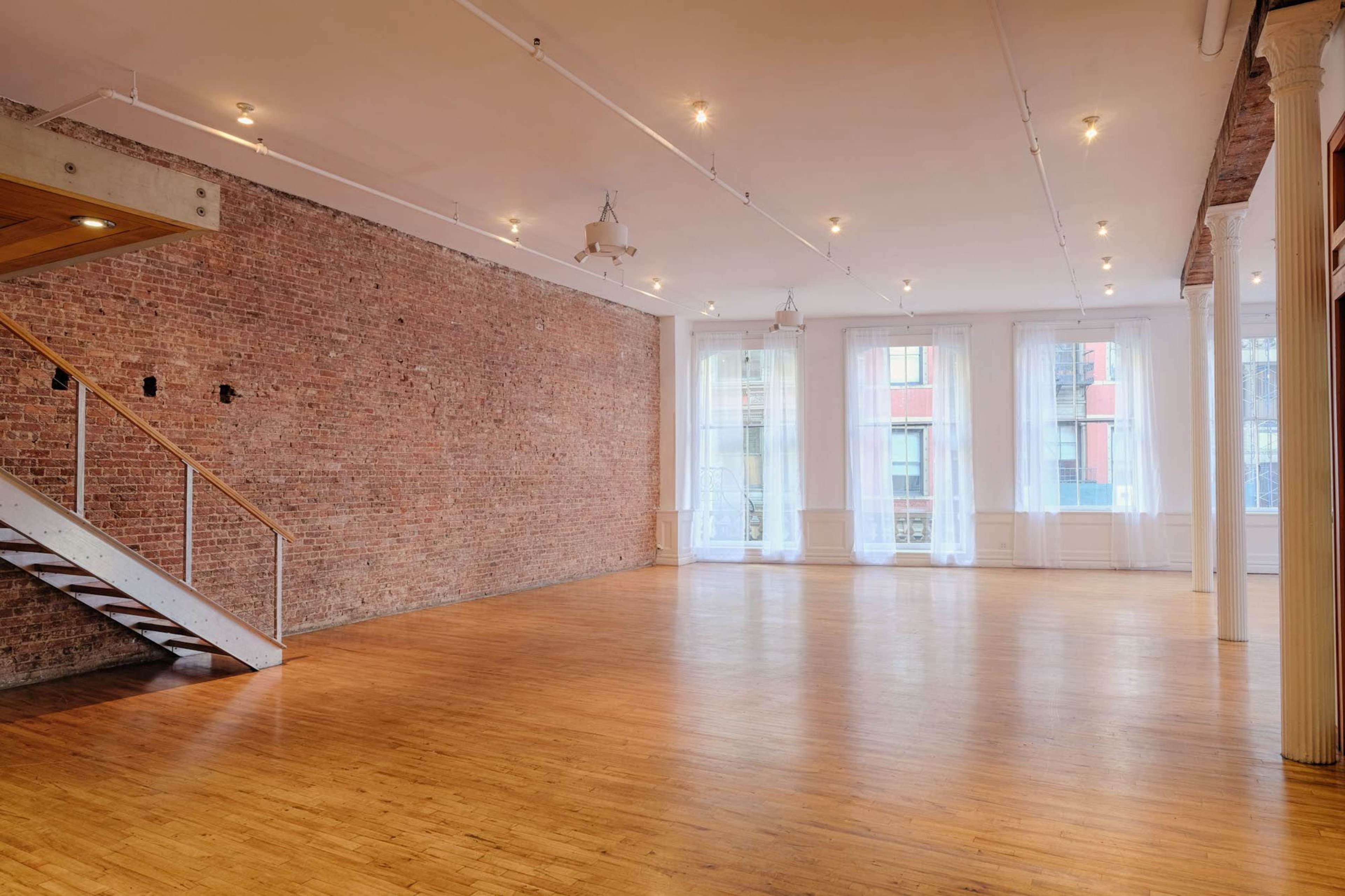 An empty, spacious room with exposed brick walls, large windows, and polished wooden floors.