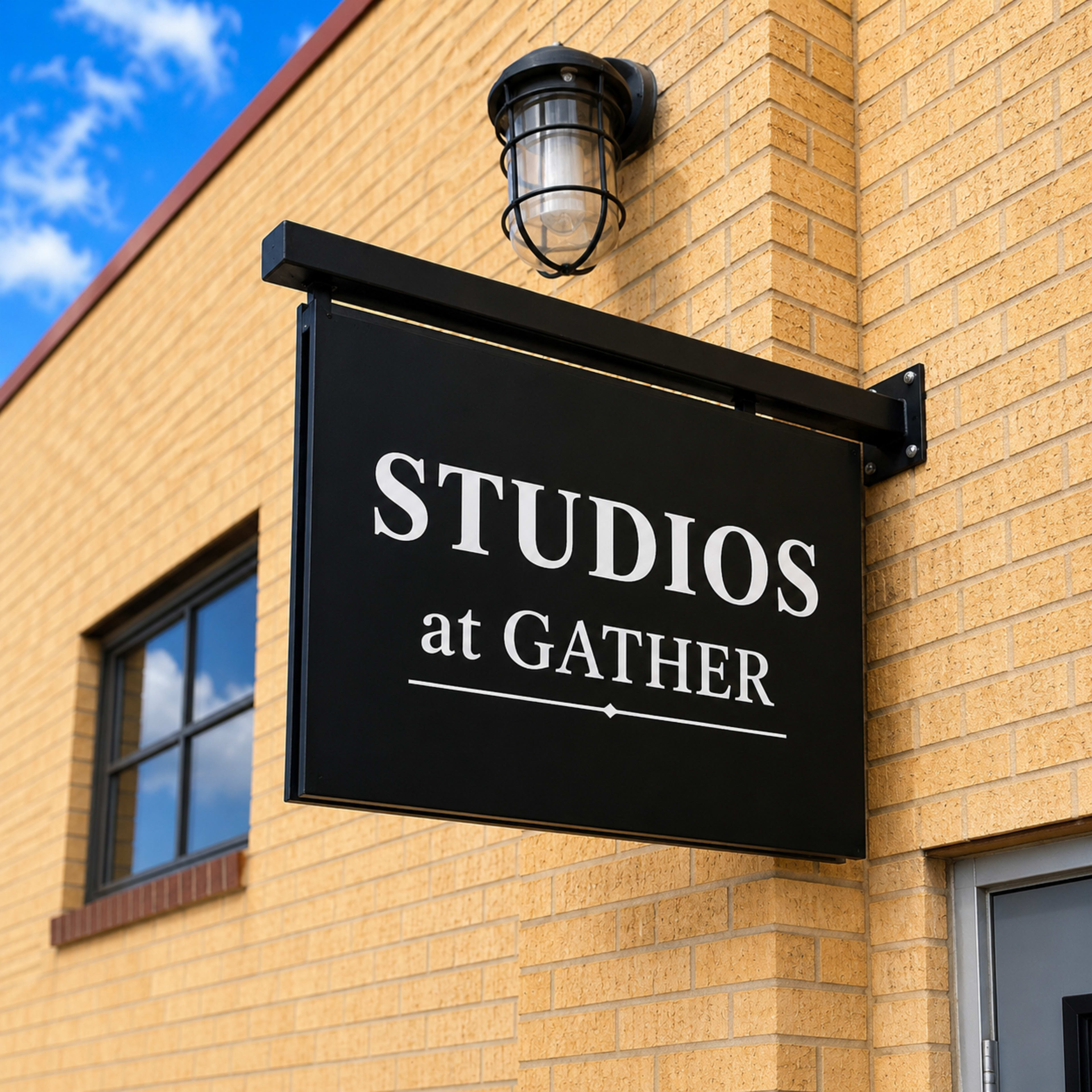 The image shows a black sign that reads "STUDIOS at GATHER" mounted on a brick building with a lamp above it.