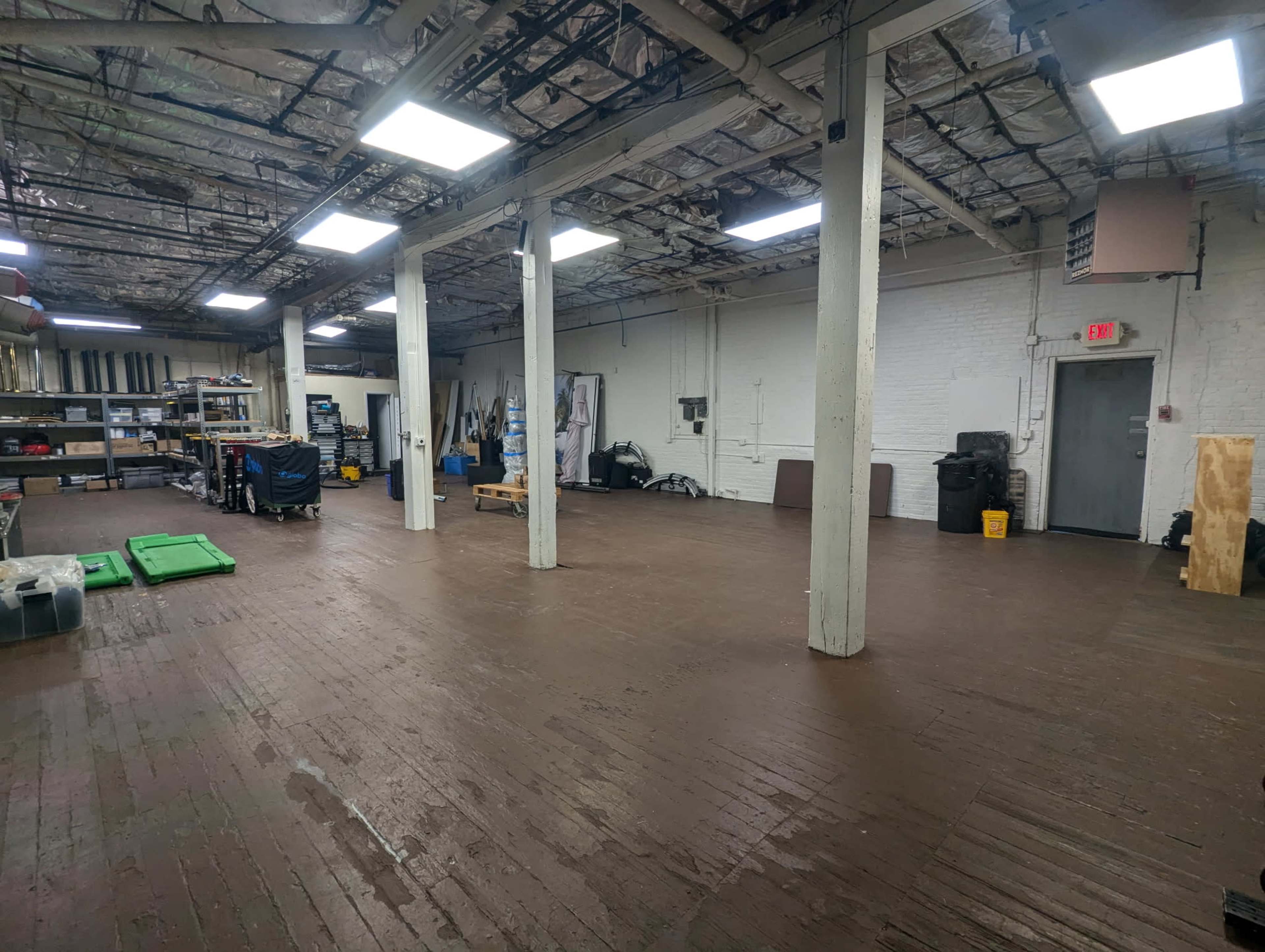 The image shows a spacious, empty warehouse with exposed ceilings, white walls, and wooden flooring, featuring several columns and some equipment along the sides.