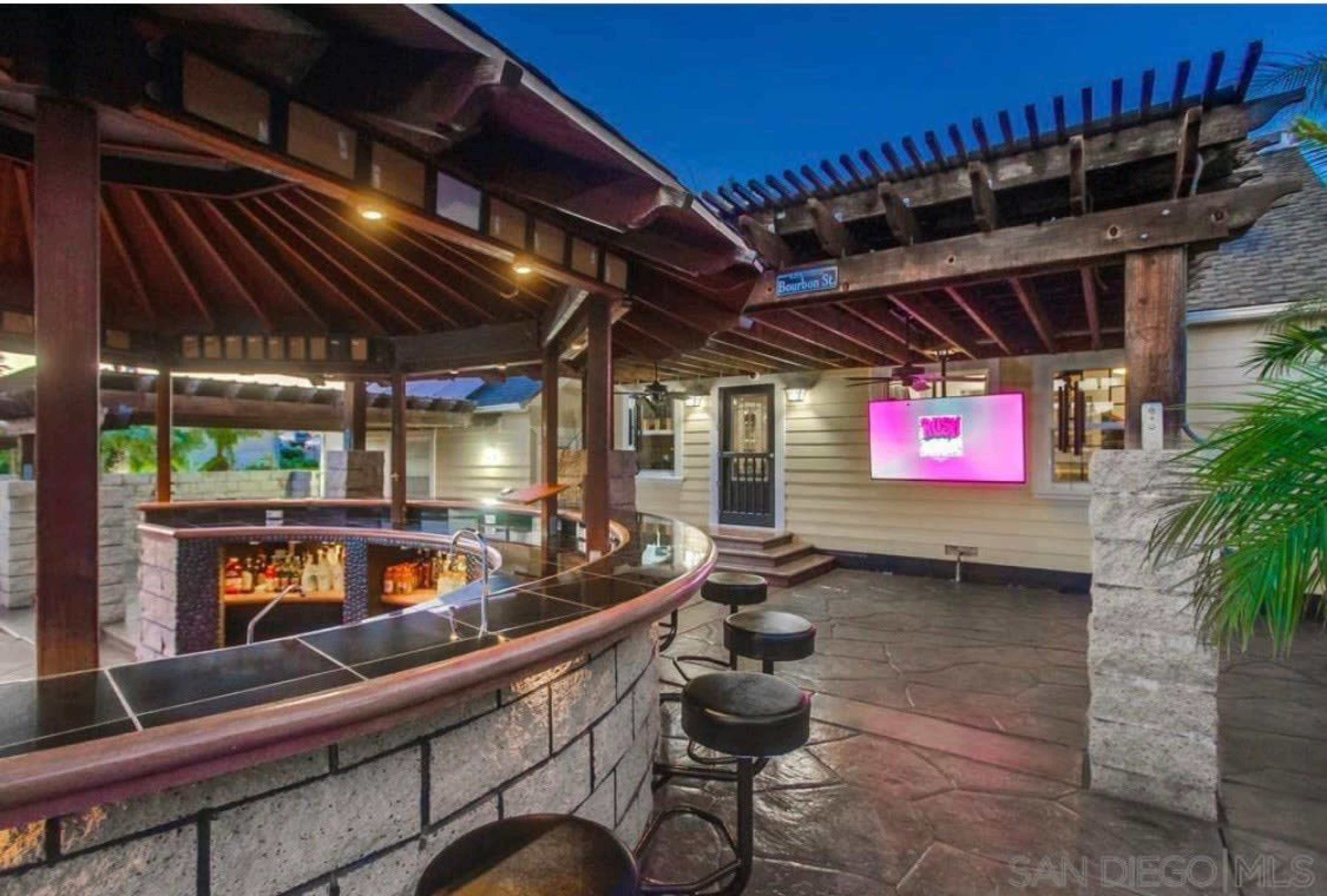 The image shows a well-designed outdoor bar area with a curved bar, stools, and a digital display under a wooden pergola.