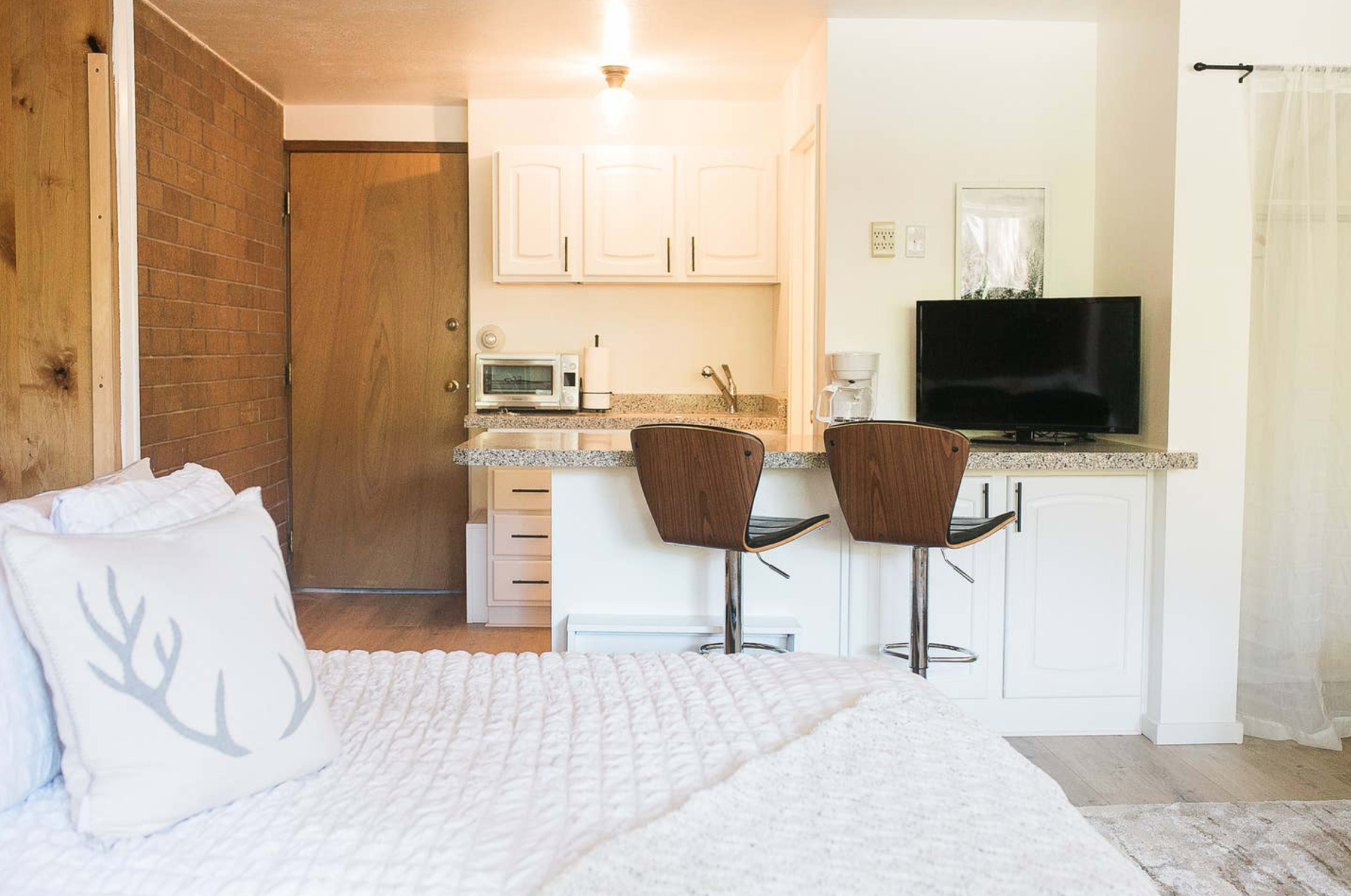 A cozy studio apartment with a bed in the foreground and a kitchen area featuring a bar with two stools and a television in the background.
