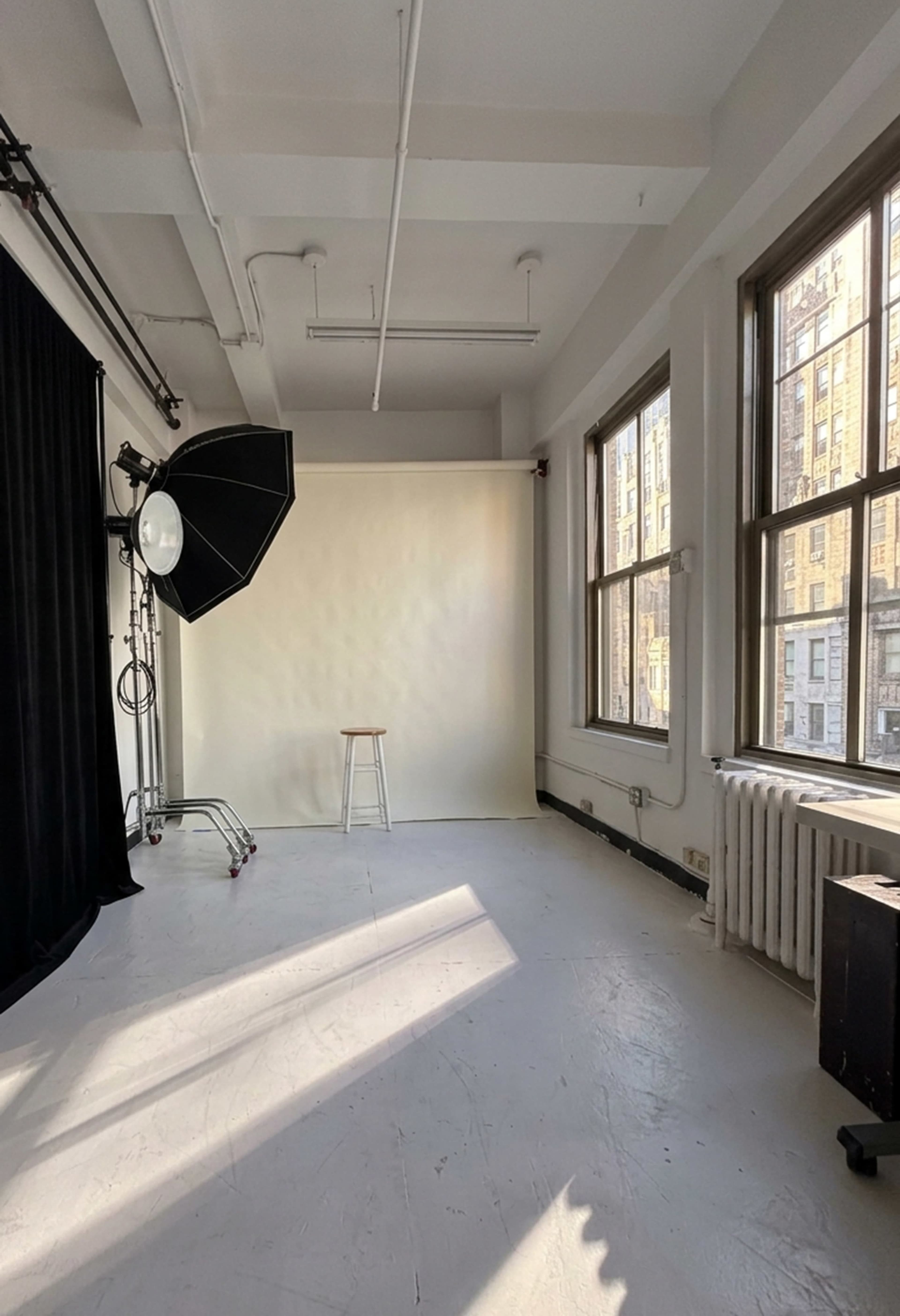 The image shows a well-lit photography studio featuring a plain backdrop, a stool, and window light streaming in through large windows.