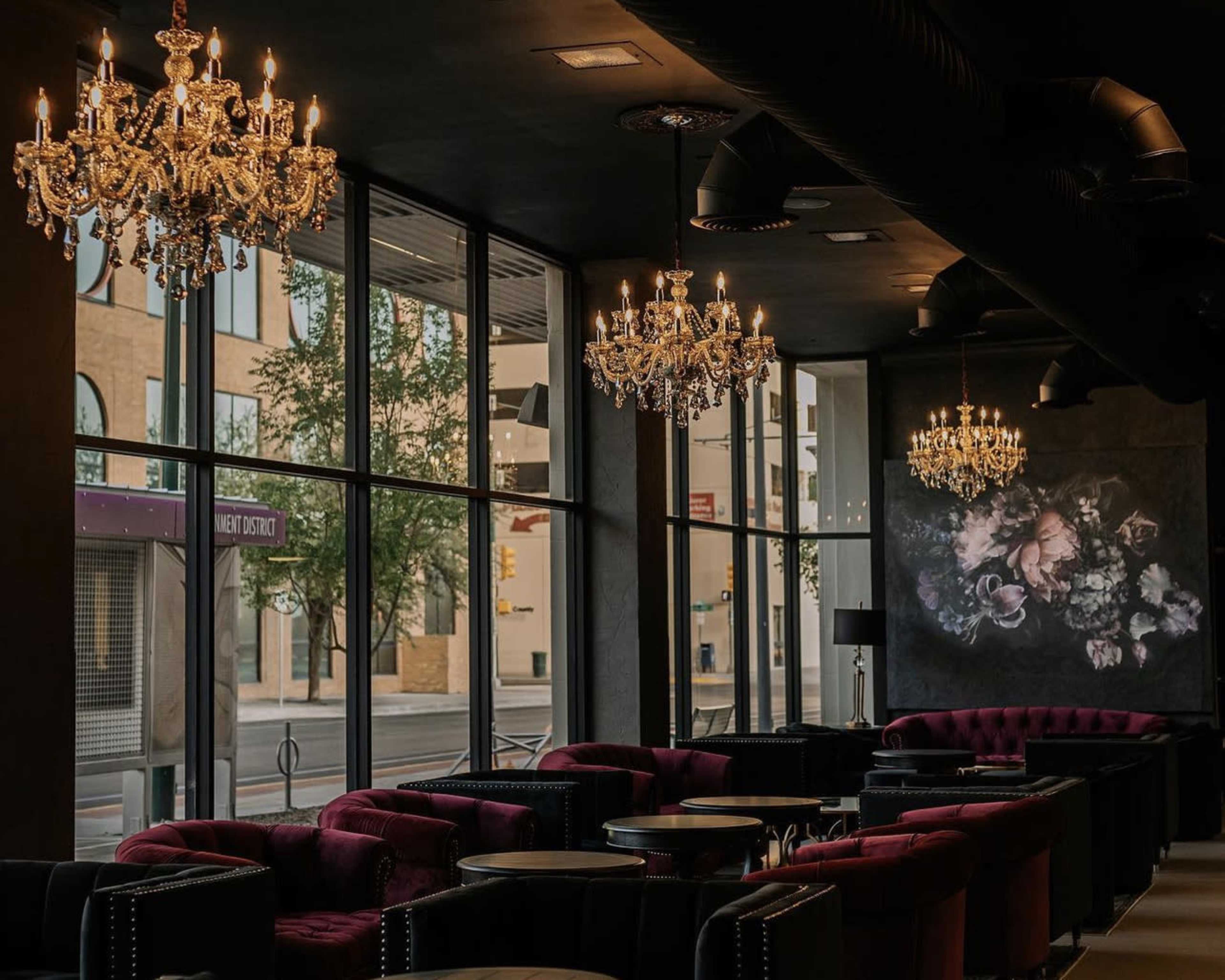A stylish lounge with velvet seating, ornate chandeliers, and large windows providing a view of the street outside.