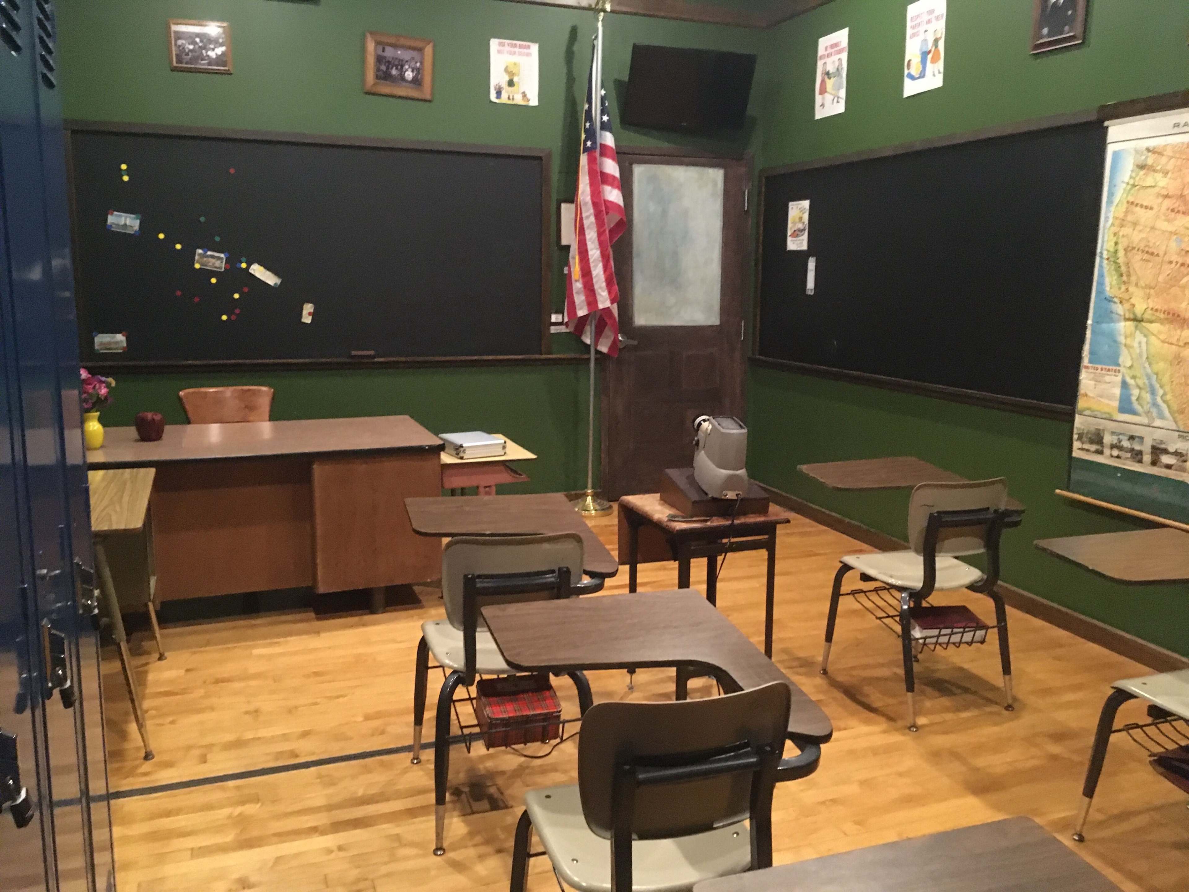 The classroom features wooden floors, a large map on the wall, and several desks arranged around a central area, with an American flag and two chalkboards in the background.