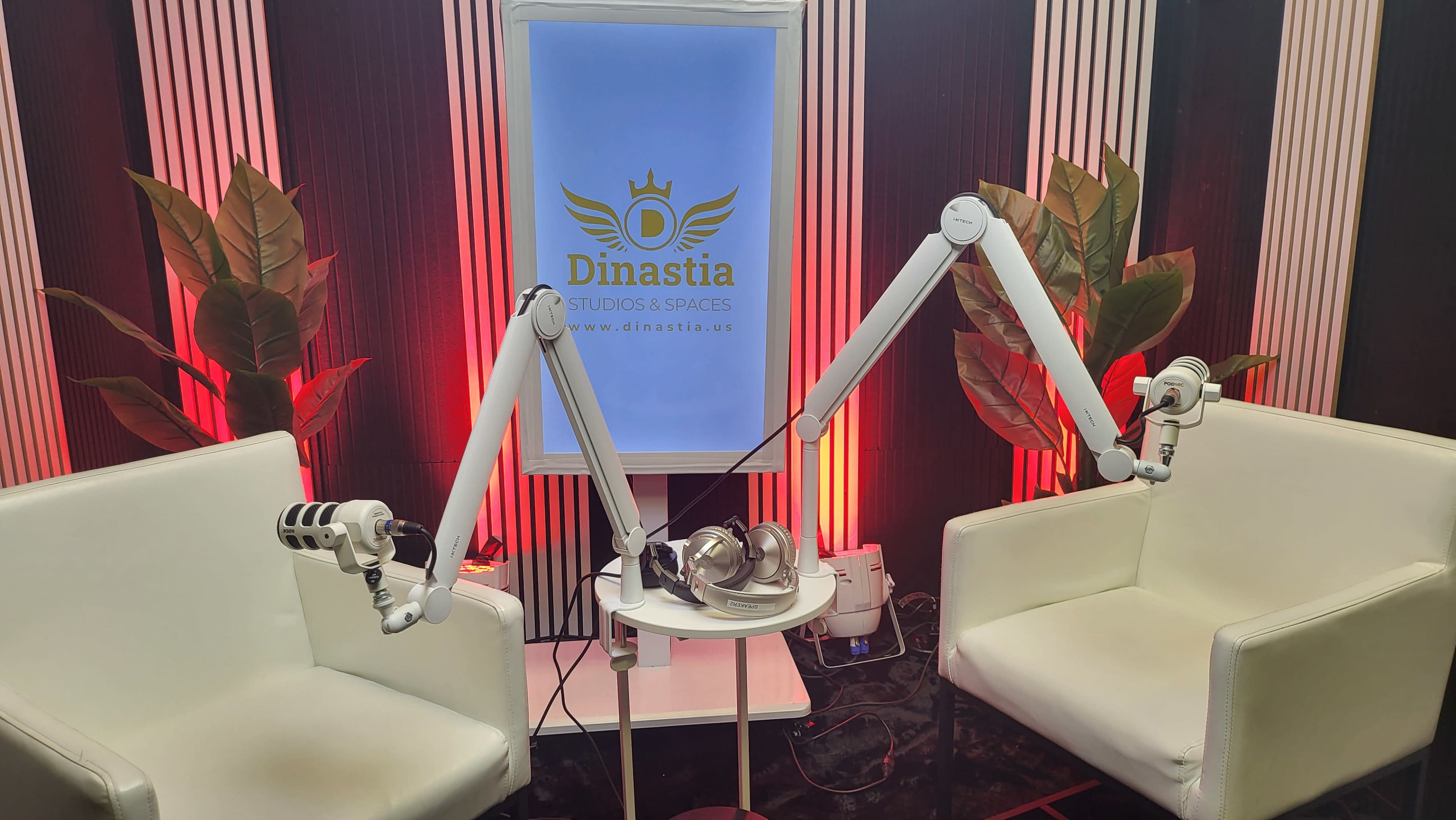 The image shows a small room with two white chairs, a round table with headphones, and two microphone stands, set against a backdrop featuring the logo of "Dinastia Studios & Spaces."