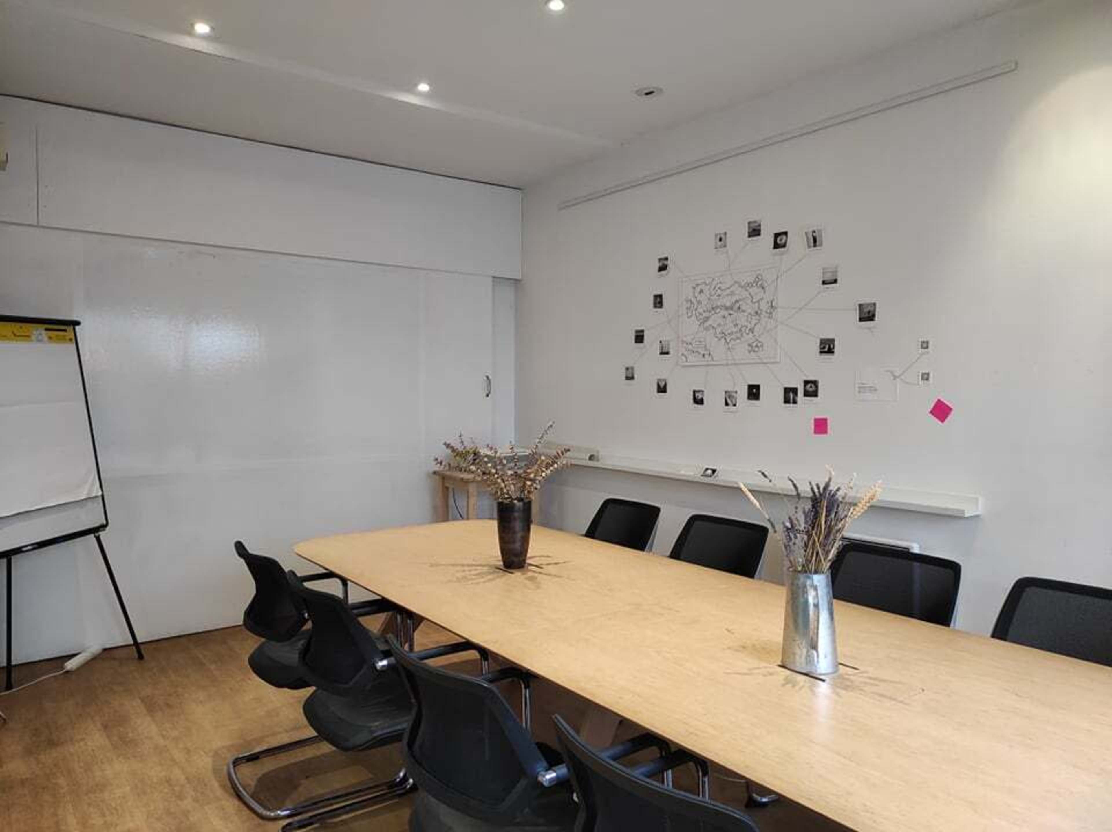 A long wooden table is set in a conference room with black chairs, a whiteboard, and various photos taped to the wall.