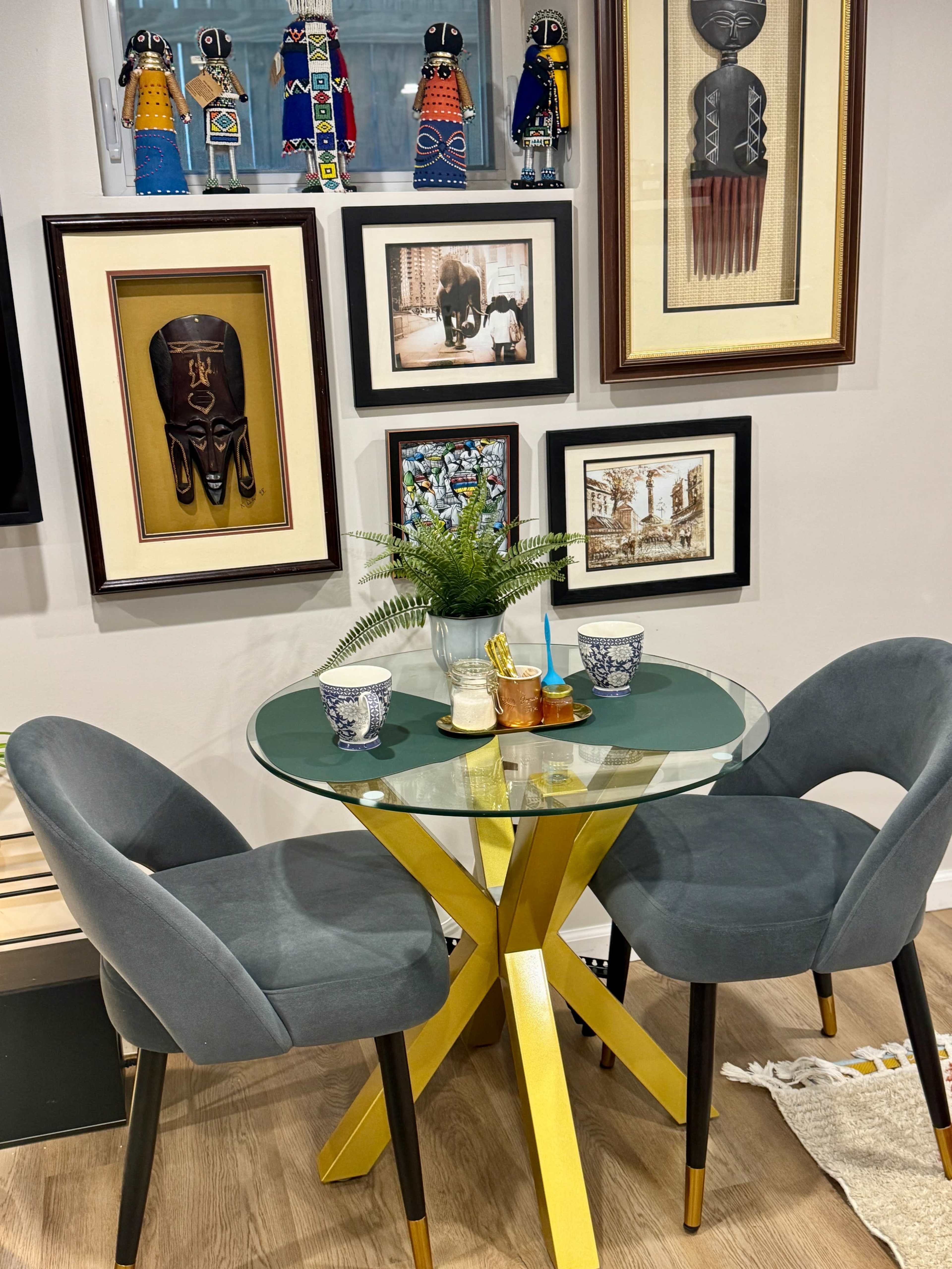 A small round glass dining table with two gray chairs is set against a wall adorned with various framed artworks and decorative figurines.