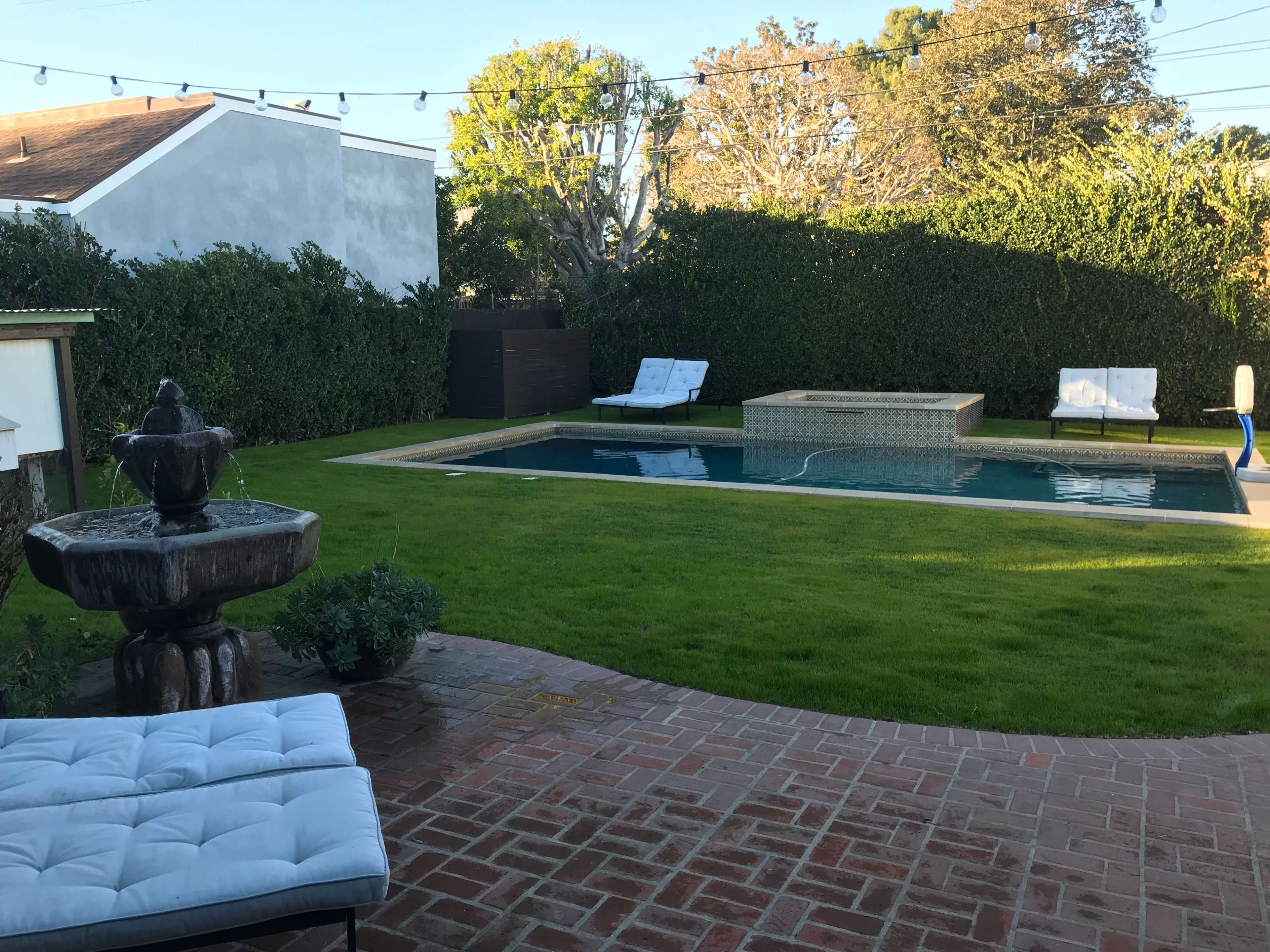 The image shows a backyard with a swimming pool, a fountain, two lounge chairs, and a well-maintained lawn surrounded by hedges.