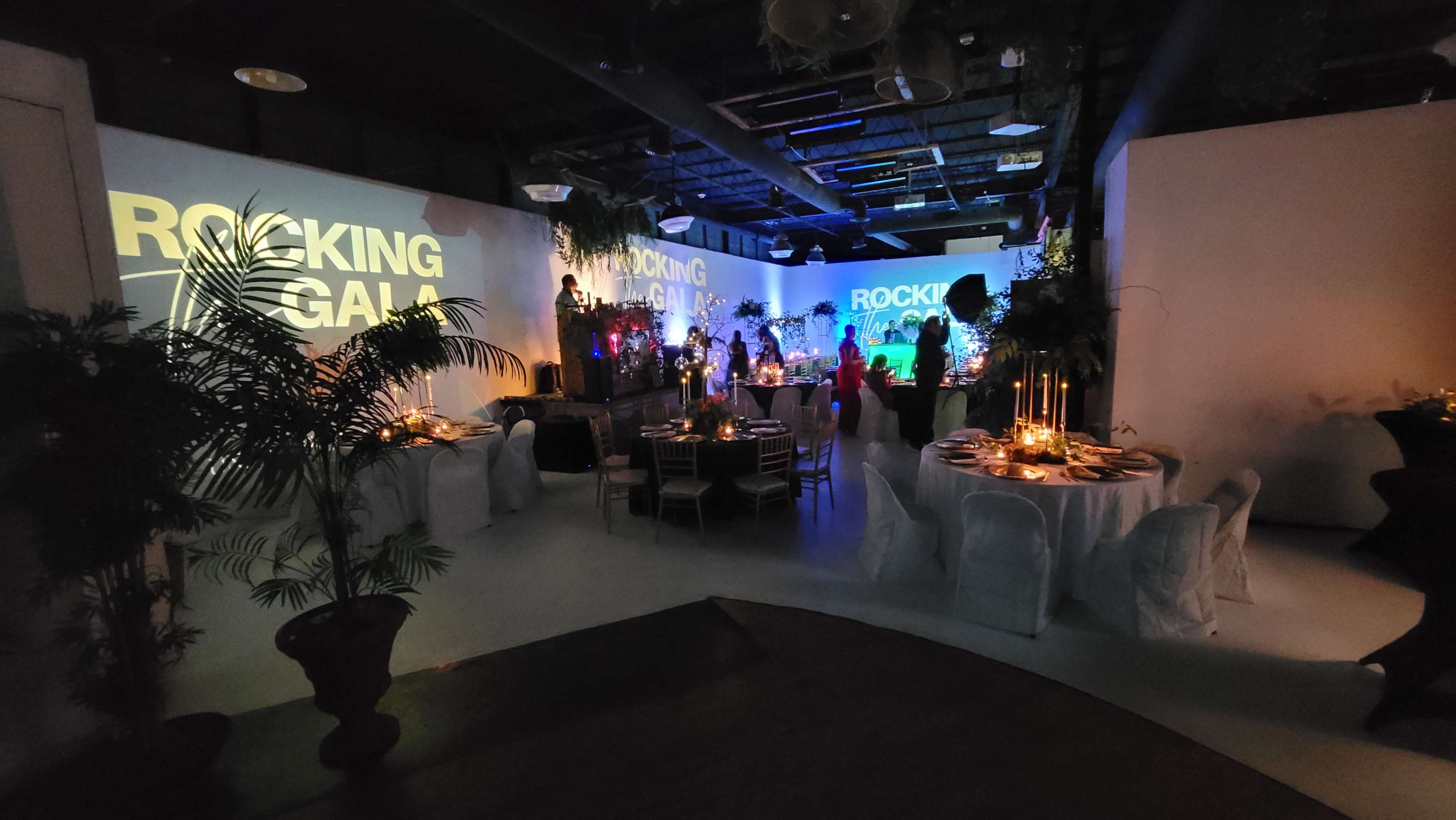 The indoor venue is set up for a gala, featuring elegantly arranged tables with candles, tropical plants, and a large projection displaying "ROCKING GALA" on the walls.