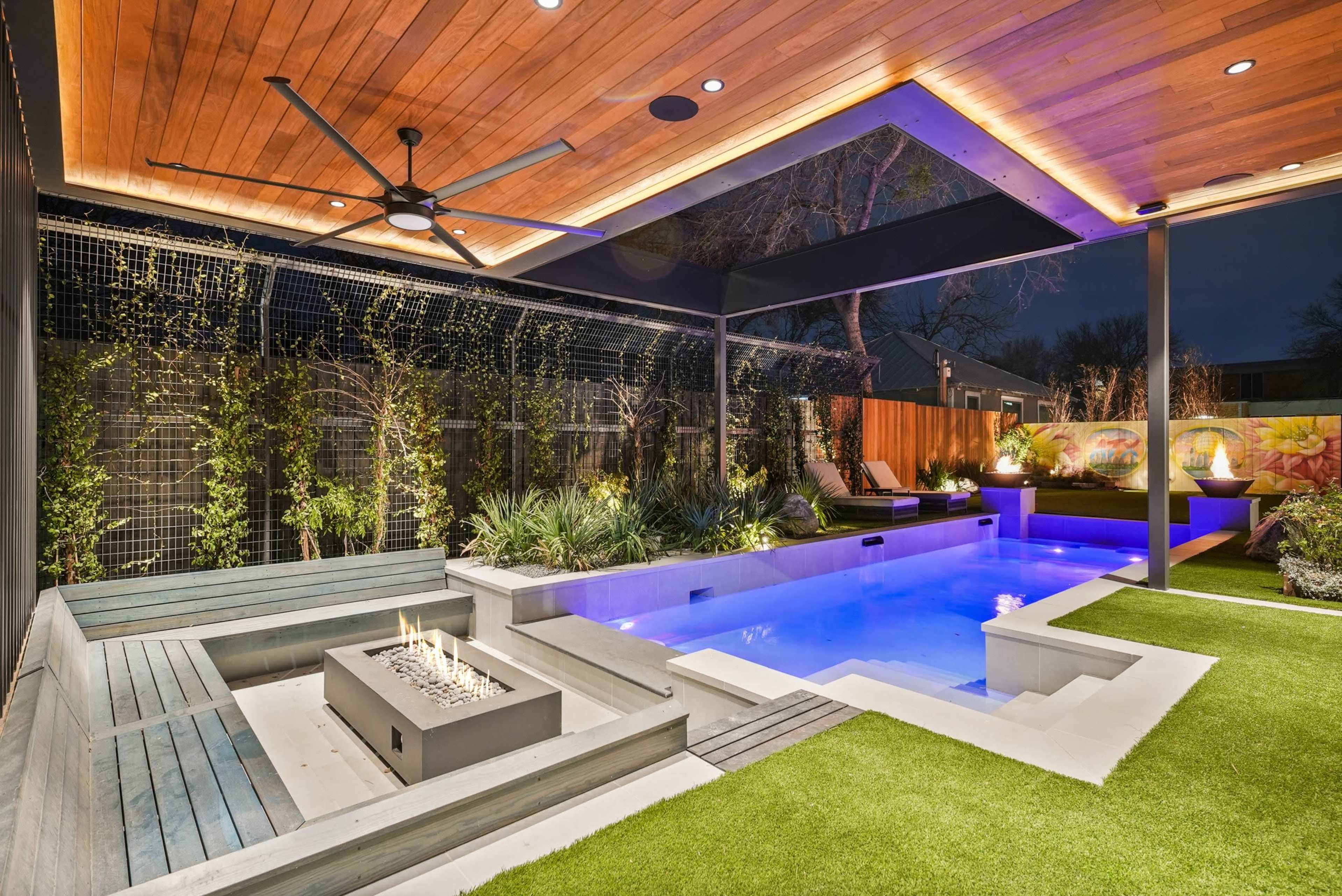 The image shows a modern outdoor space featuring a rectangular swimming pool, surrounded by grass and patio seating, with a covered area overhead that includes a fan and accent lighting.