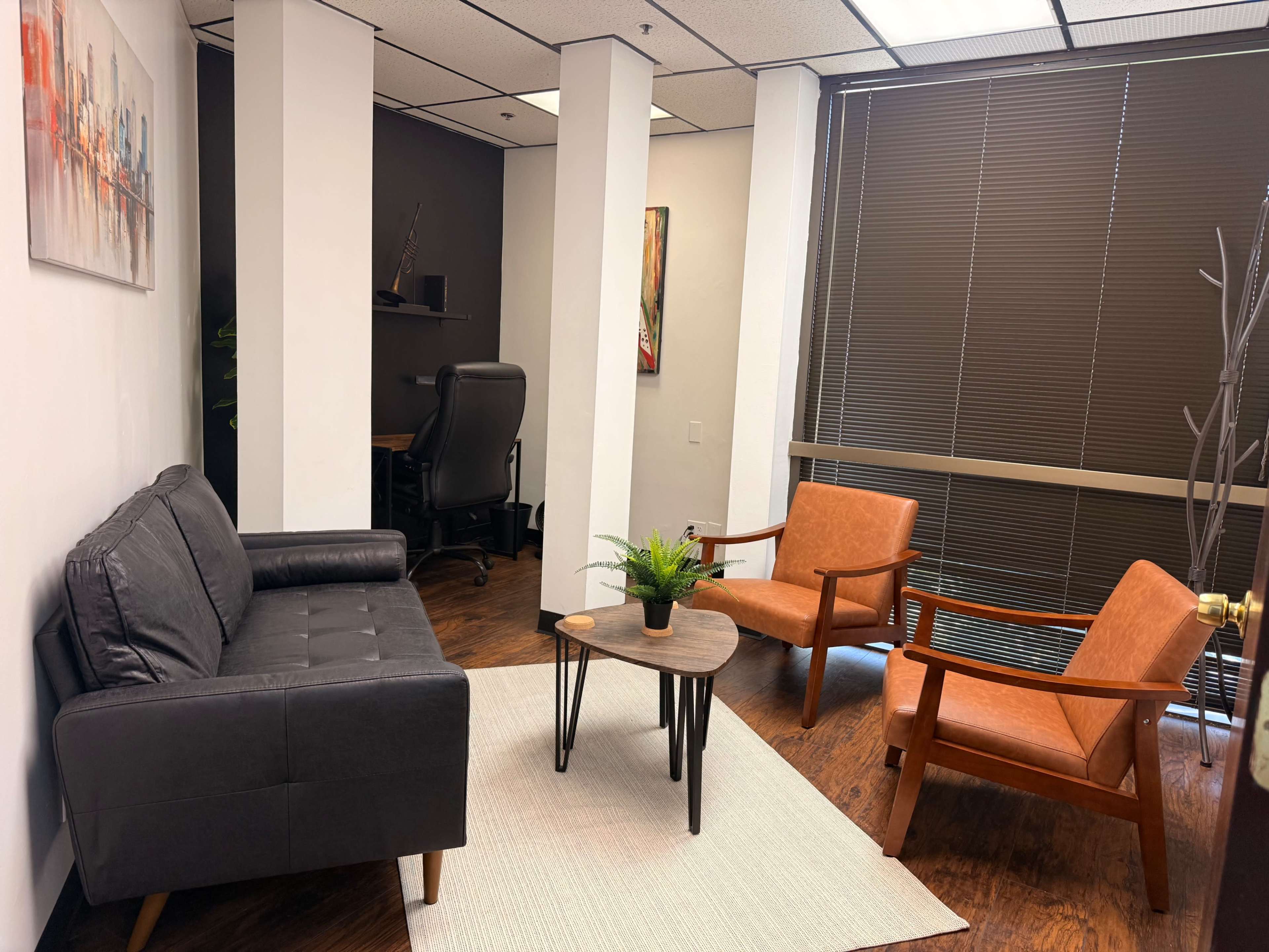 The image shows a small office waiting area with a black sofa, two orange chairs, a round coffee table, and a potted plant on a light rug beneath overhead fluorescent lights.