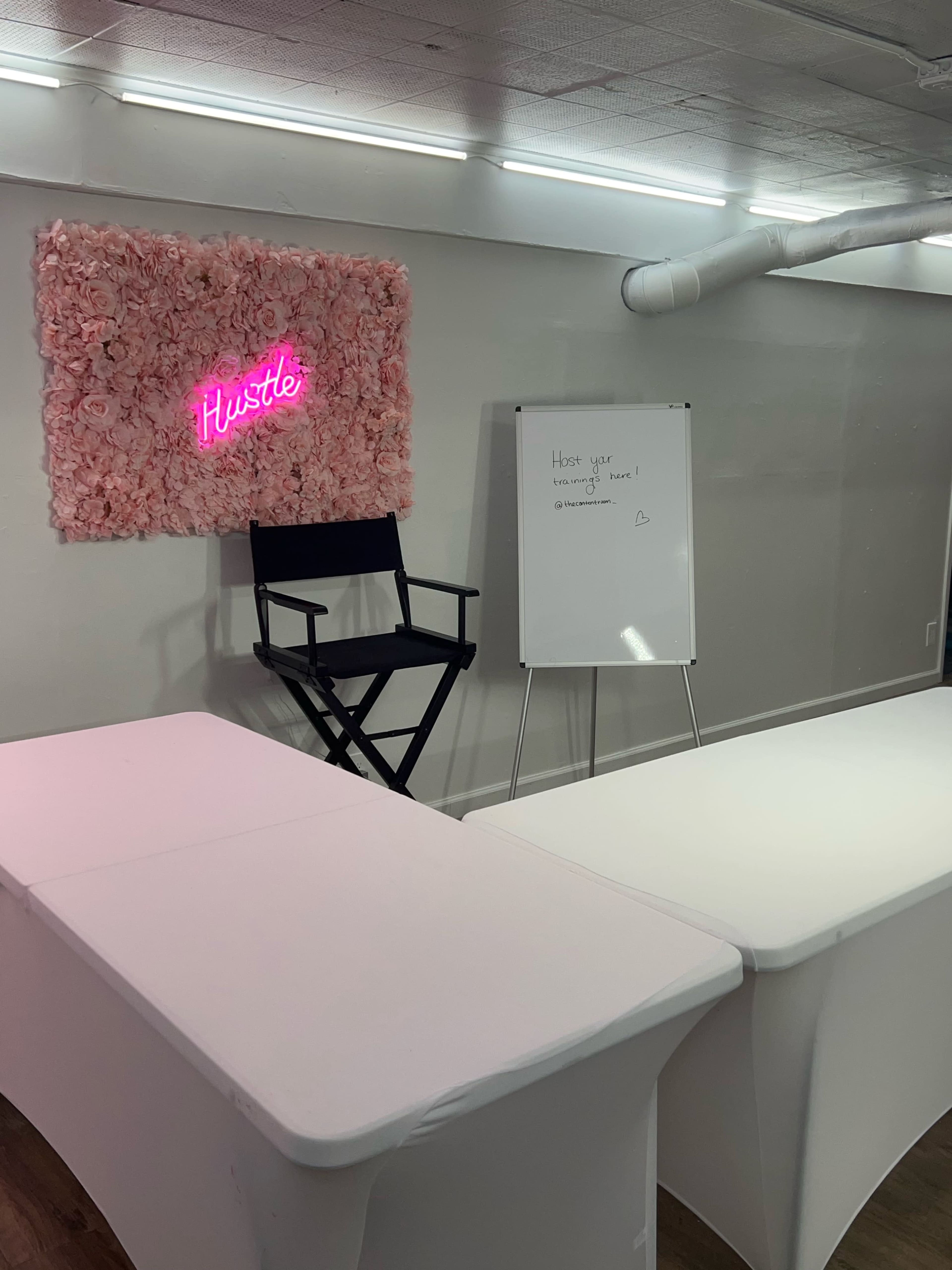 A minimalist workspace featuring a floral wall with a neon sign that reads "Hustle," a director's chair, and a whiteboard.