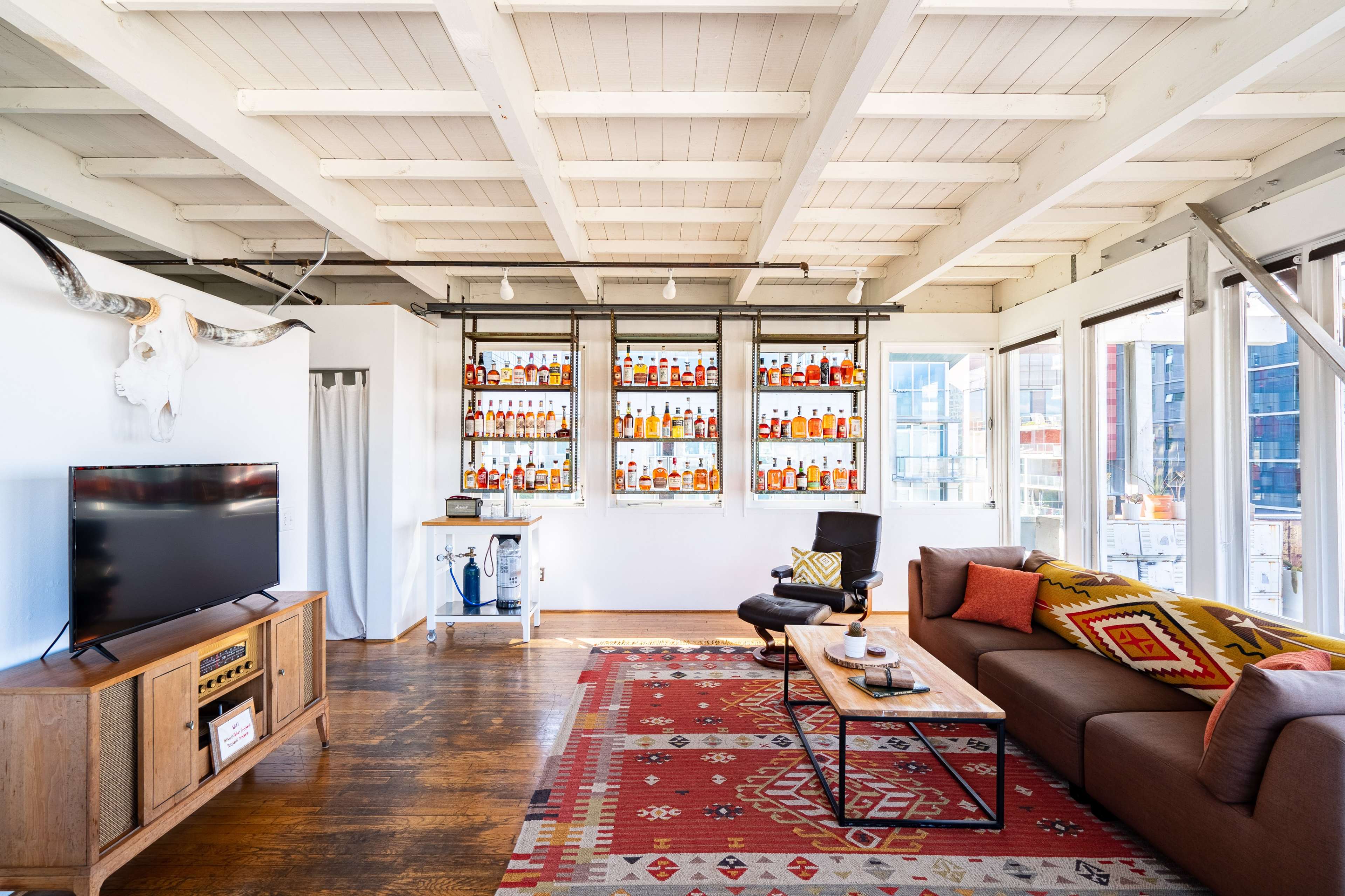The image shows a modern living room with wooden floors, a television, and a decorative shelf filled with bottles, set against large windows.
