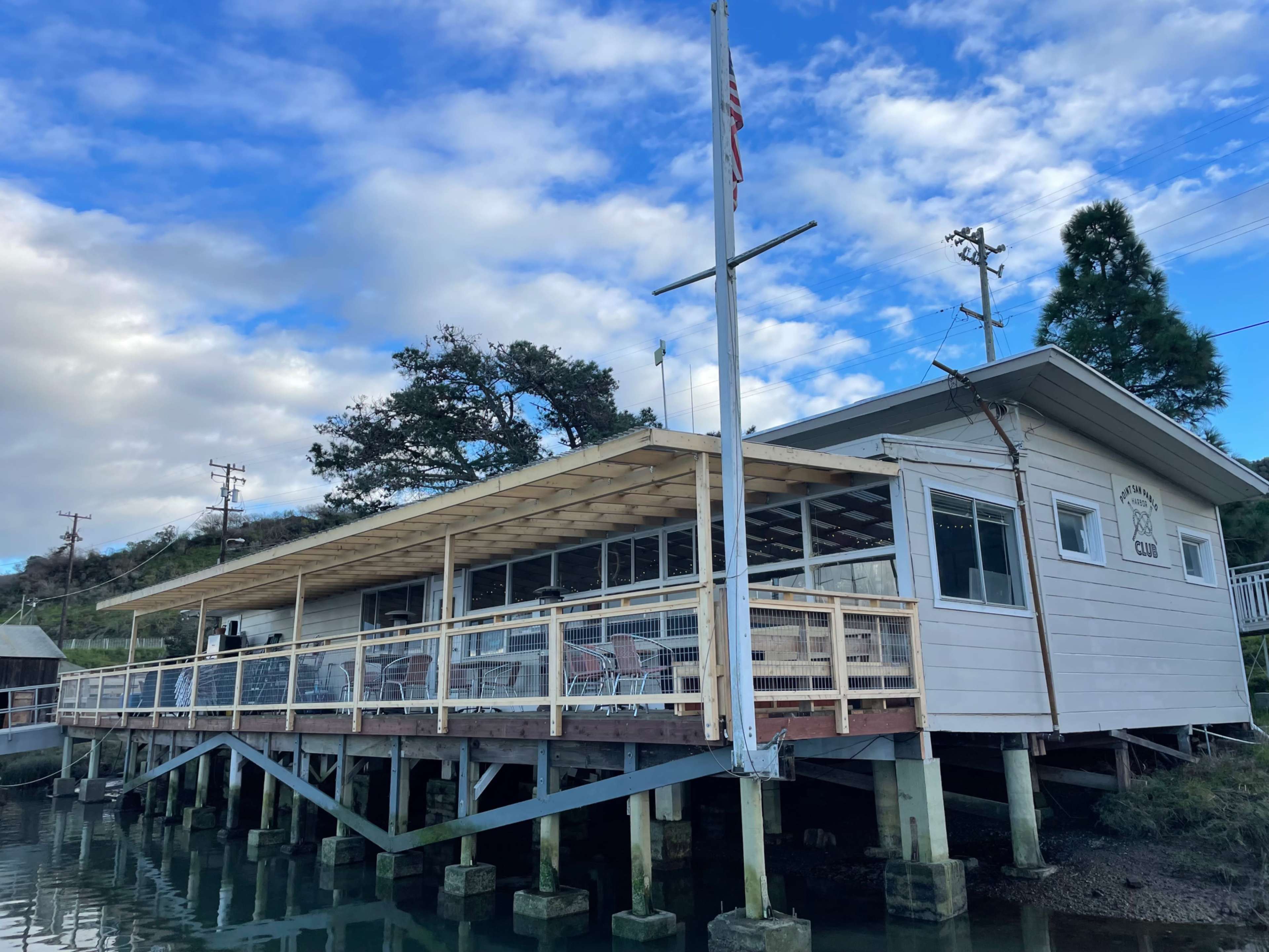 The building is a wooden structure on stilts overlooking a body of water, featuring a deck with chairs and a flagpole.