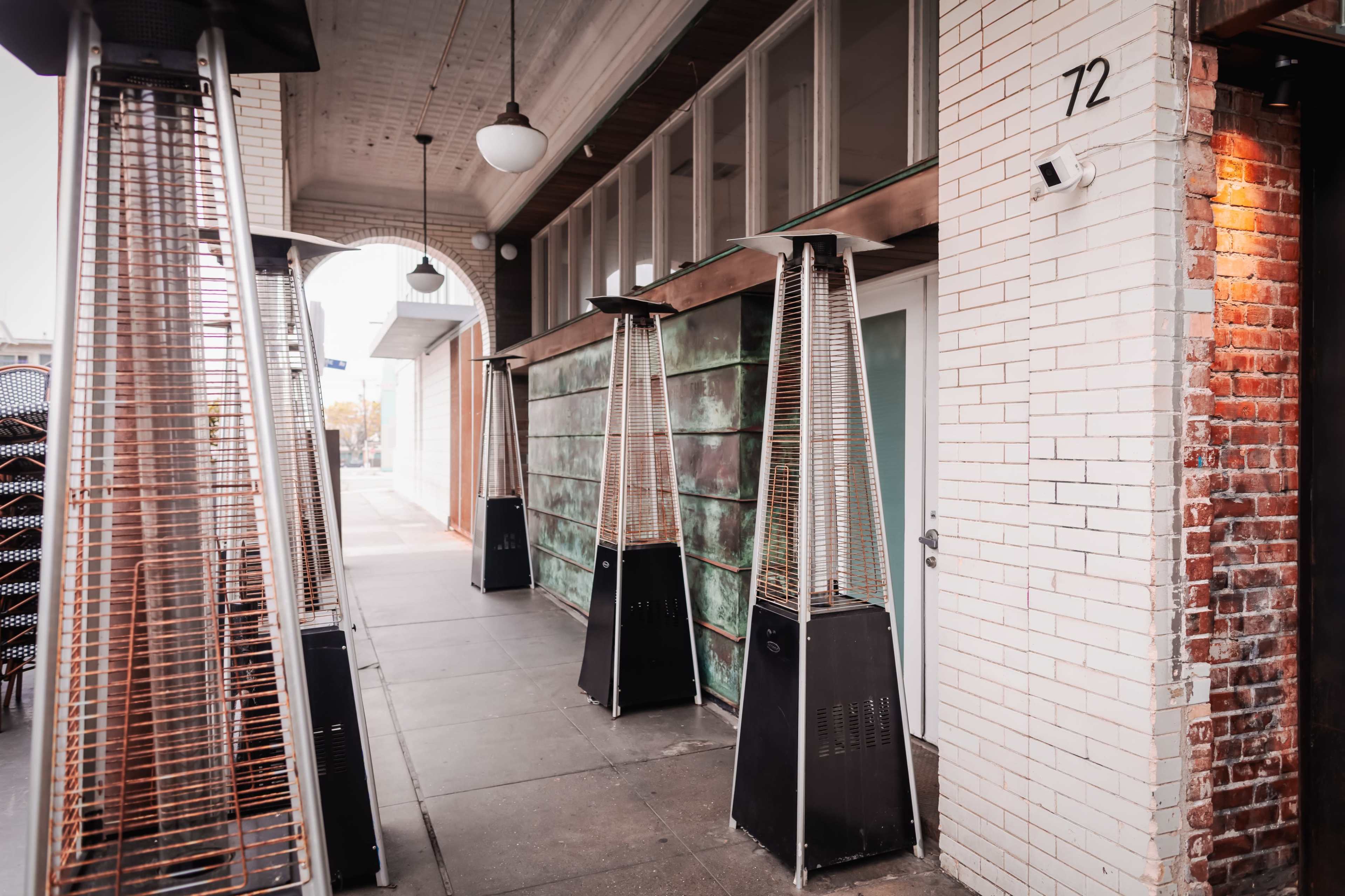 The image shows several tall outdoor heaters lined up along a sidewalk in front of a building with a weathered metal wall.
