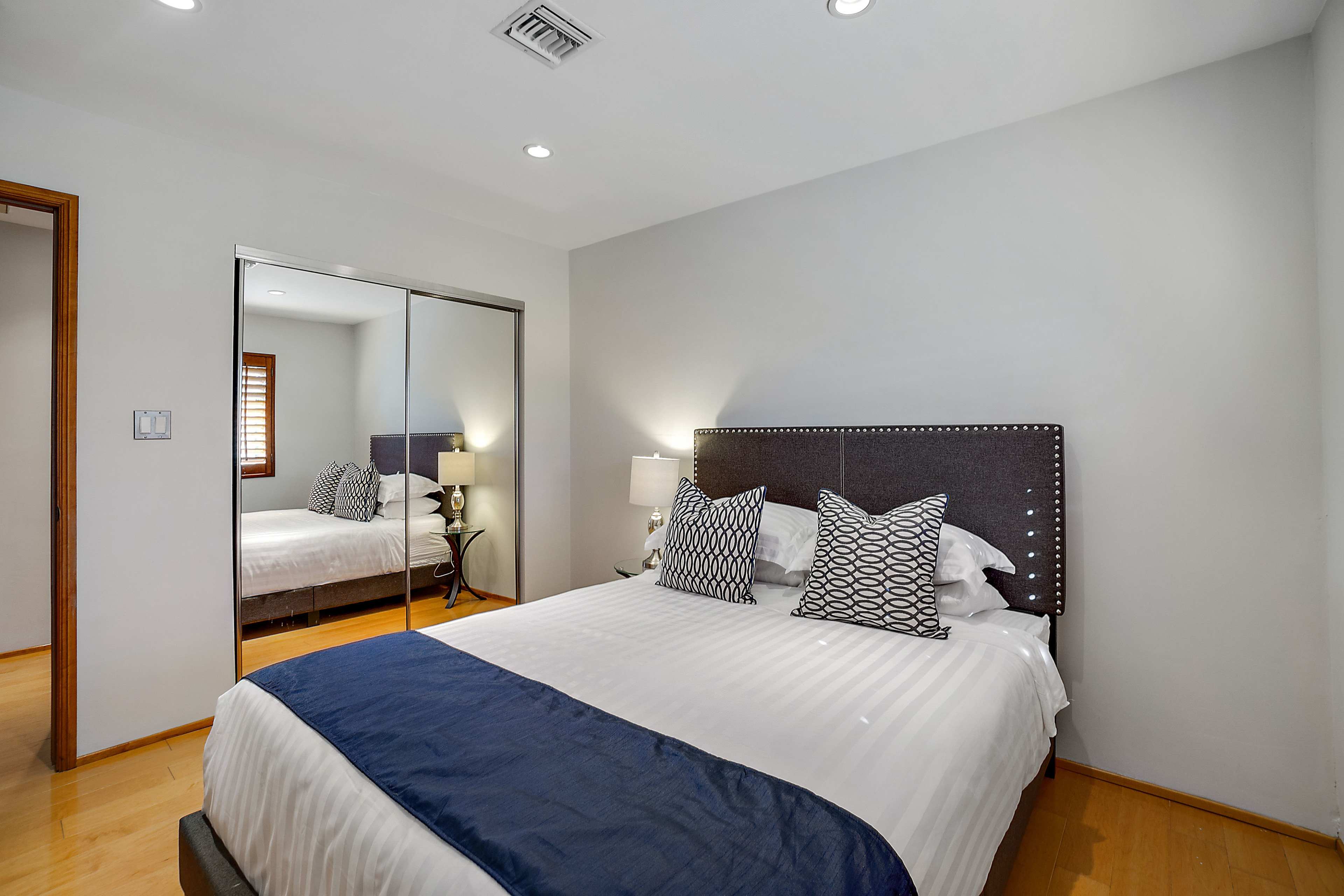 A well-lit bedroom featuring a bed with patterned pillows, a bedside table, and a mirrored closet.