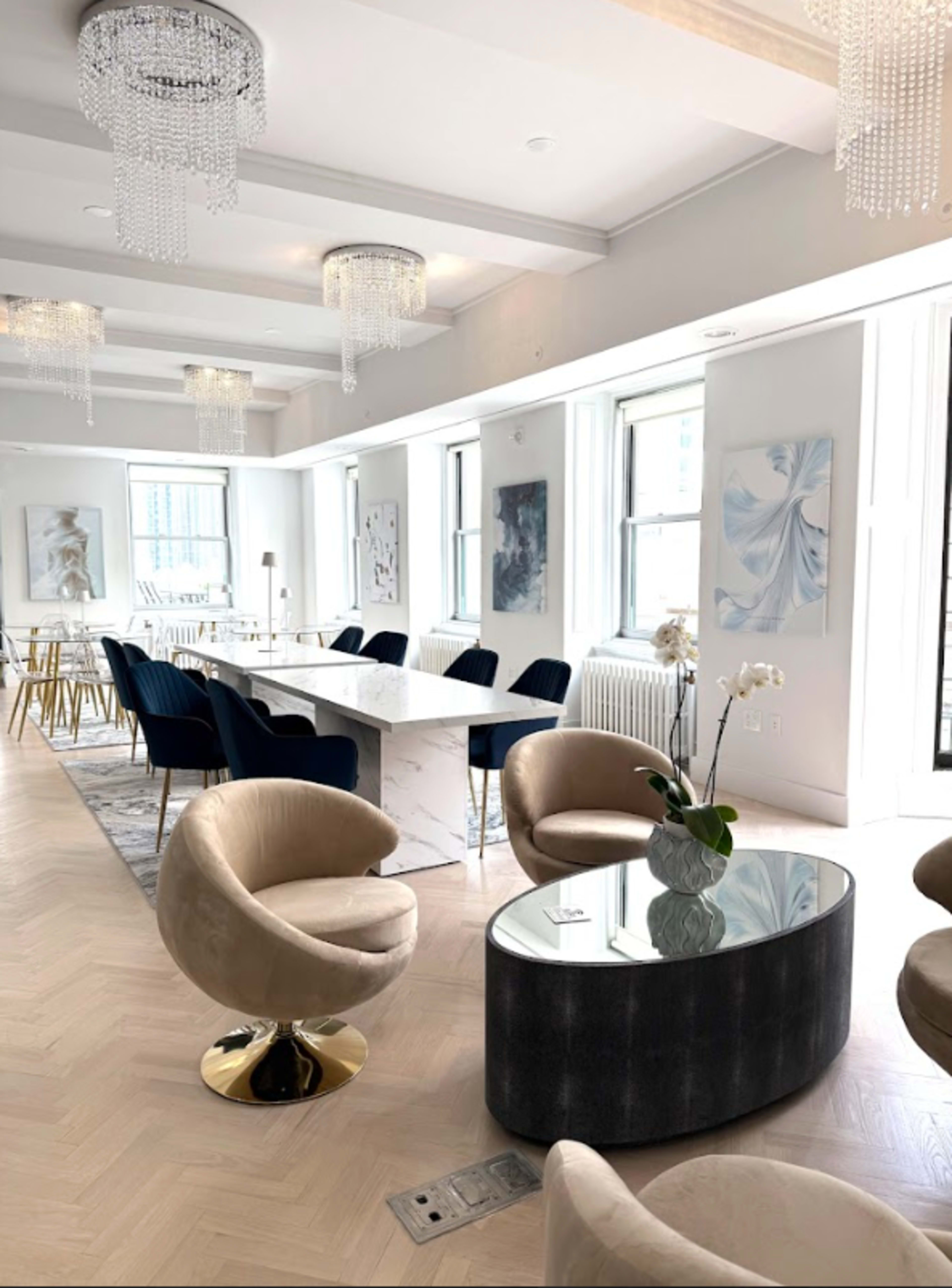 The image features a modern, well-lit interior with plush seating, a dining table, and elegant chandeliers.