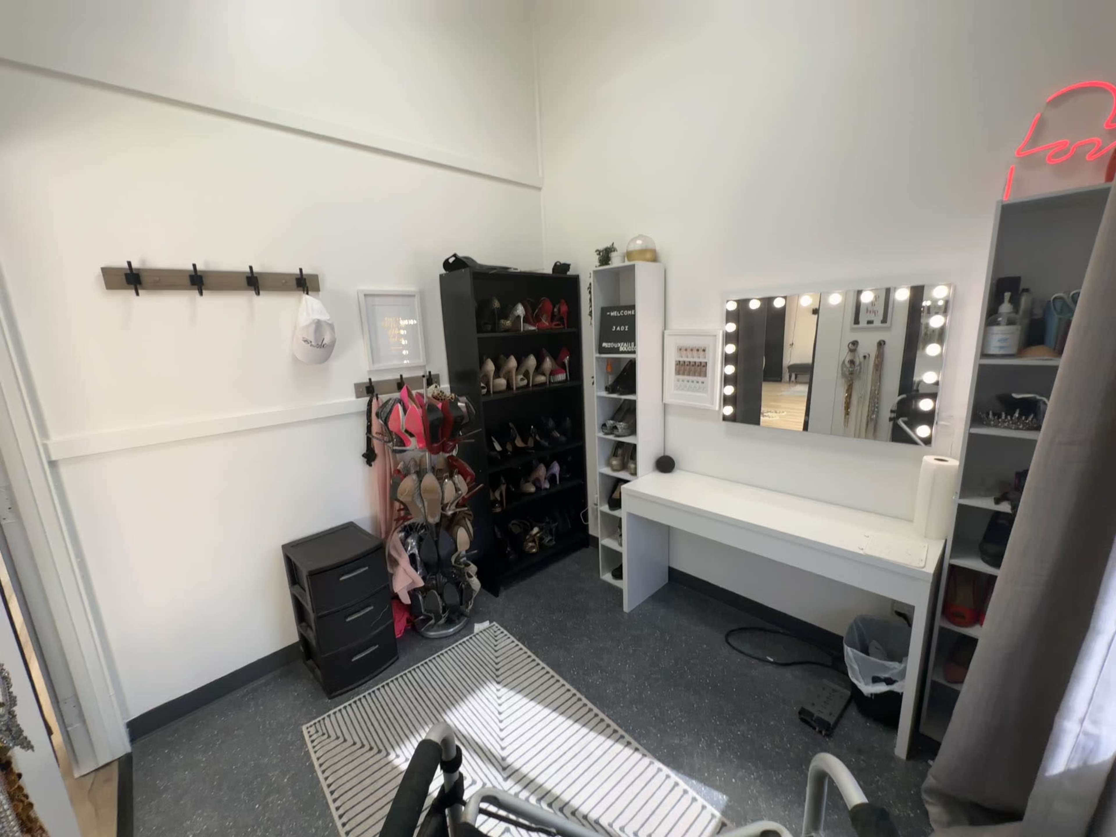 A small room featuring a makeup vanity with illuminated mirrors, a shoe rack filled with high heels, and a clothes rack along the wall.