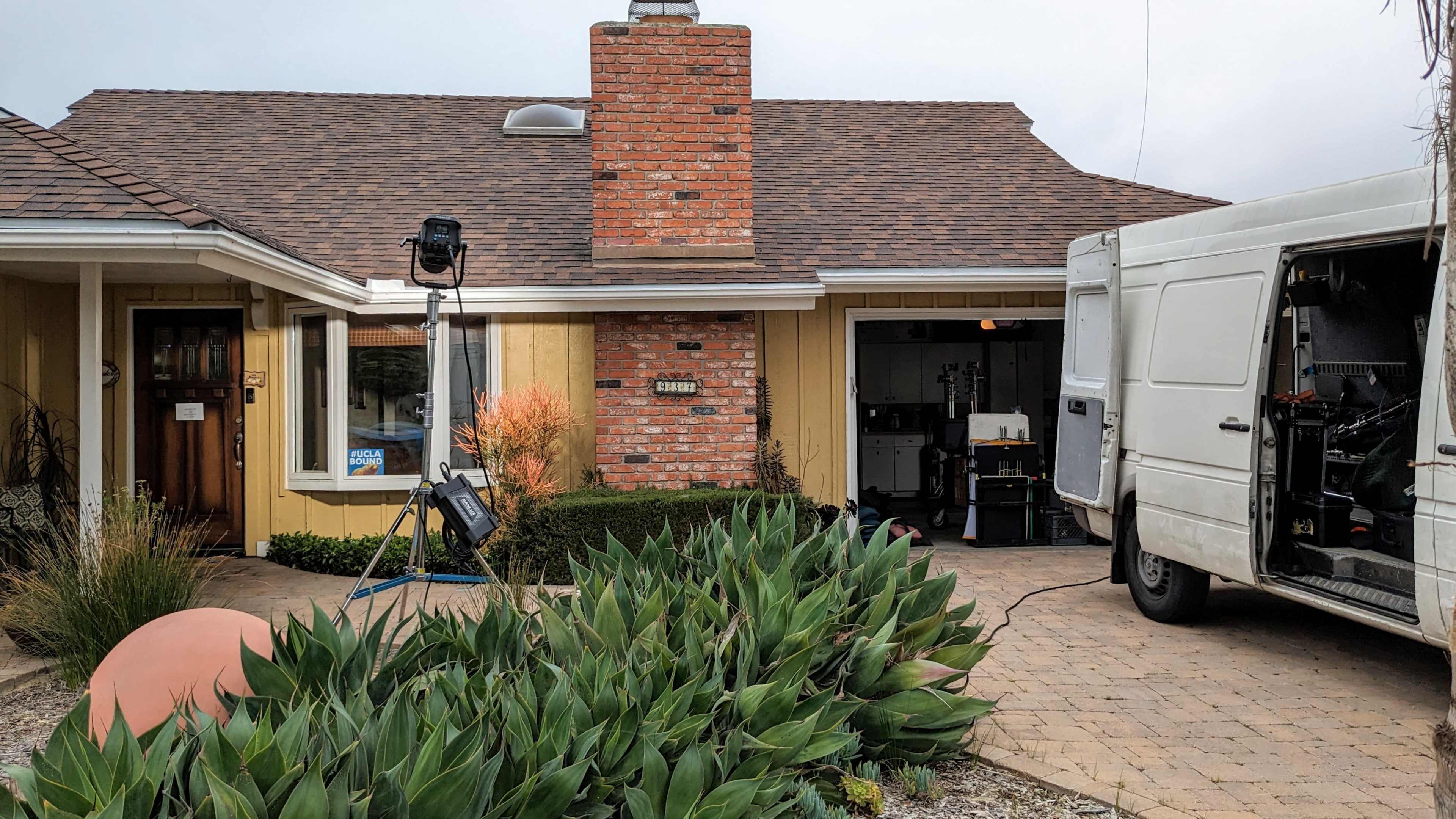 A van is parked next to a house with a red brick chimney, and a lighting setup is positioned on the front lawn.