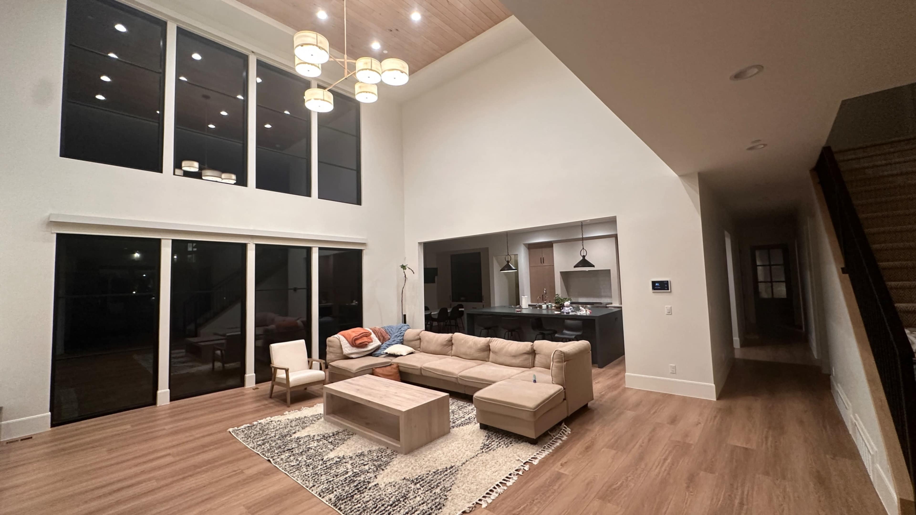 A spacious living room with a sectional sofa, coffee table, and large windows that illuminate the area, adjacent to a kitchen with an open layout.