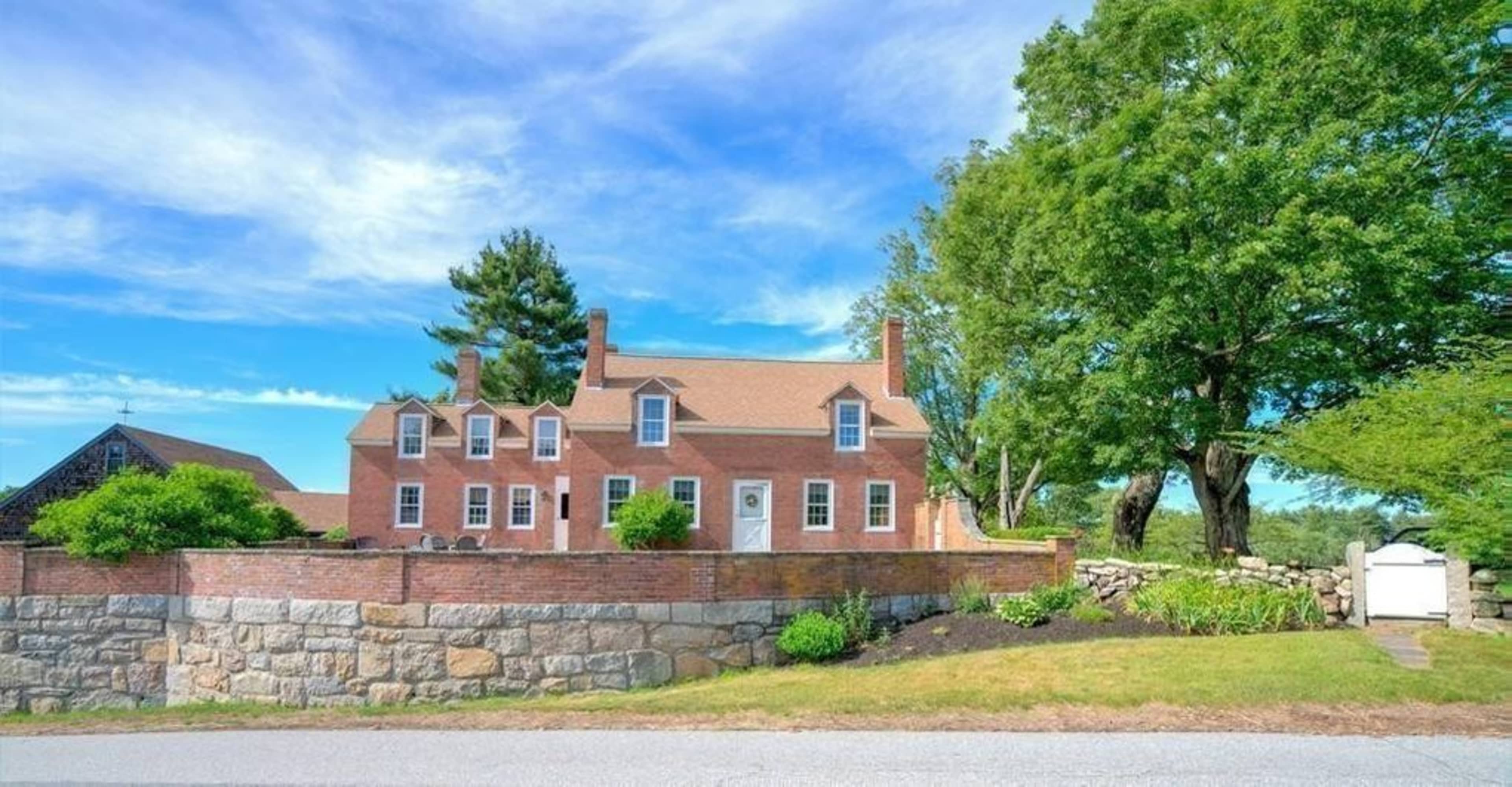 A brick house with a gabled roof is surrounded by a stone wall and lush greenery.