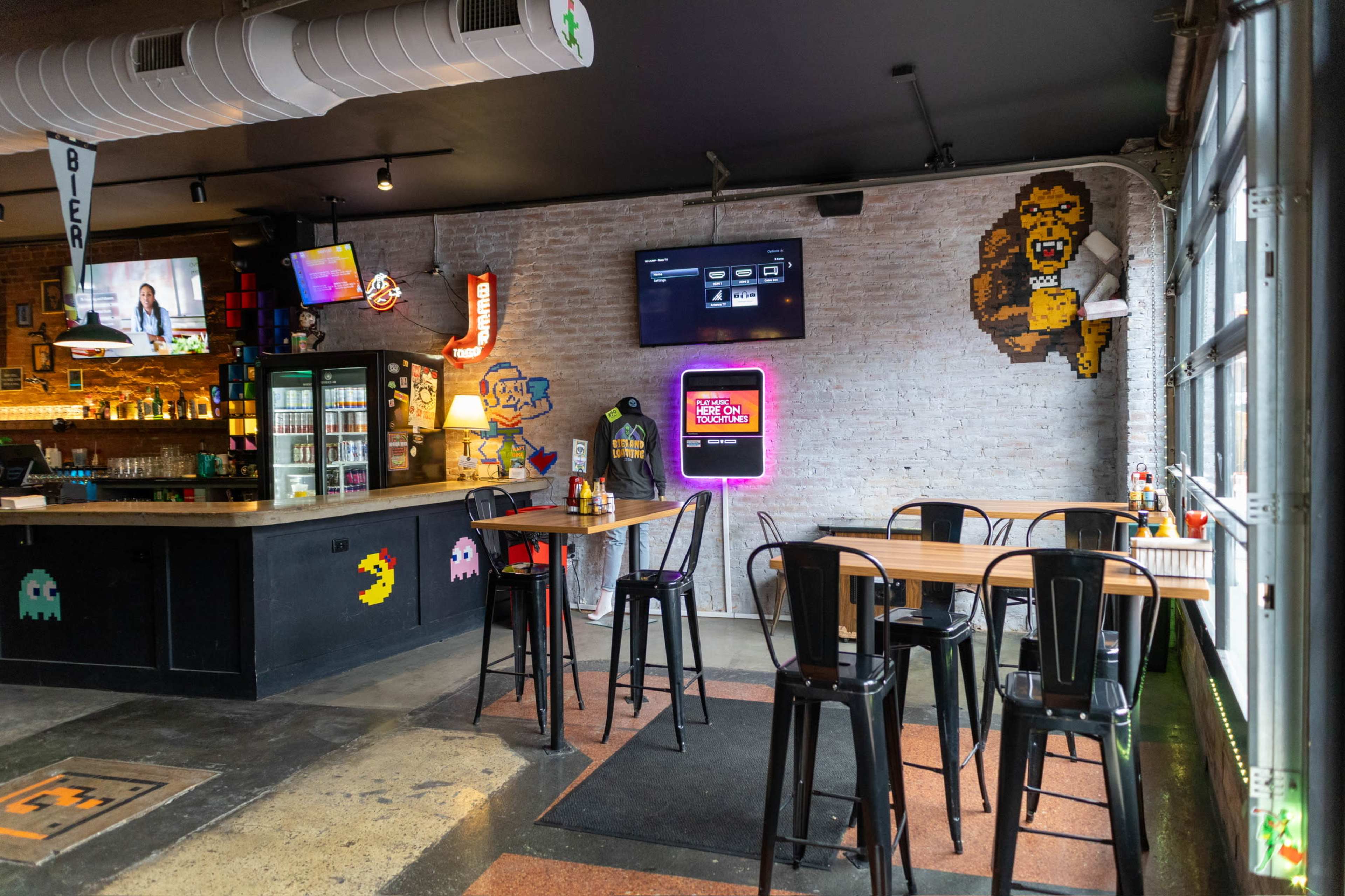 The image shows a modern bar interior with high tables, black chairs, and a retro video game theme, featuring pixel art characters on the walls.