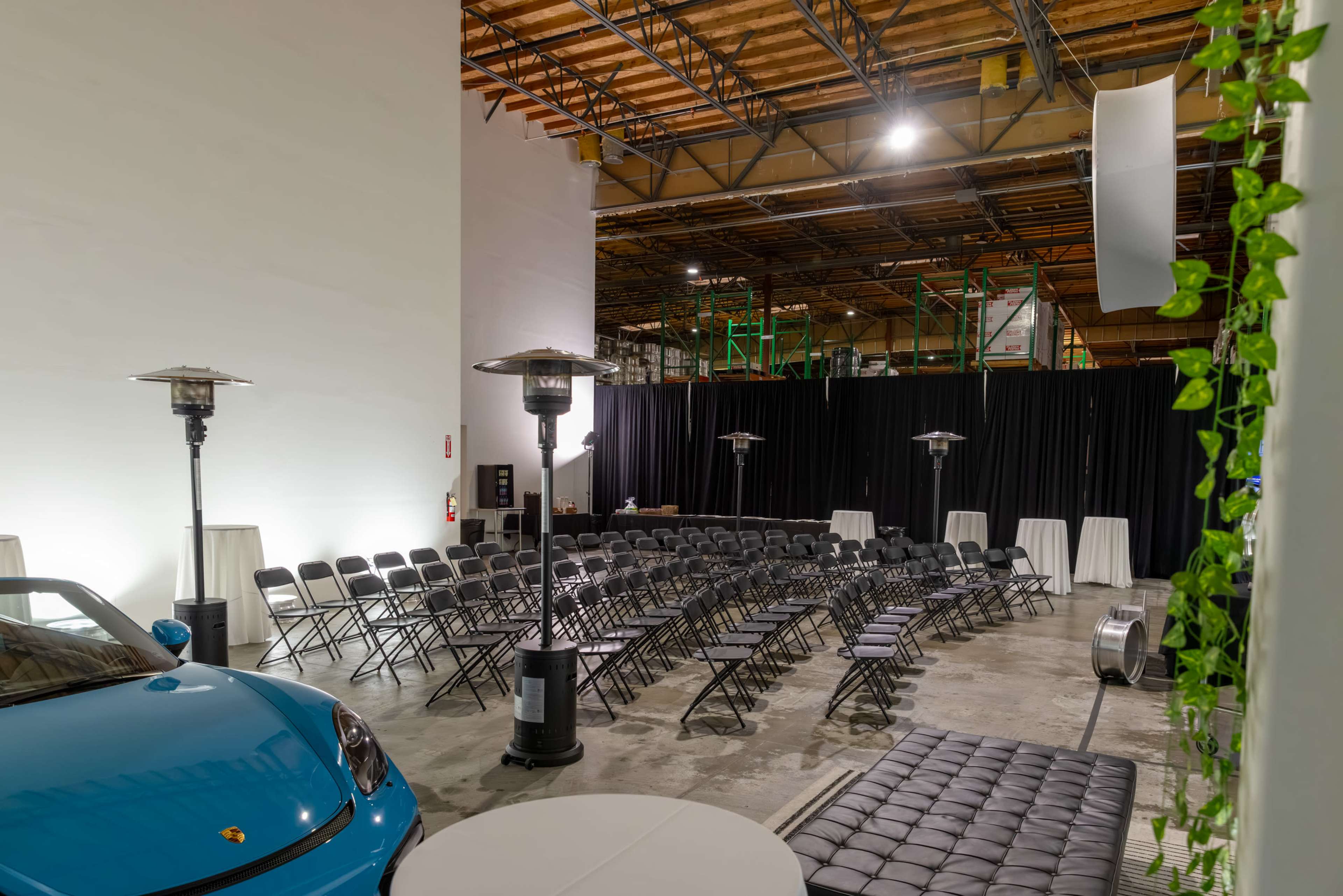 The image depicts an indoor event space with rows of chairs arranged in a semicircle, a blue car parked nearby, and standing heaters set up along the sides.