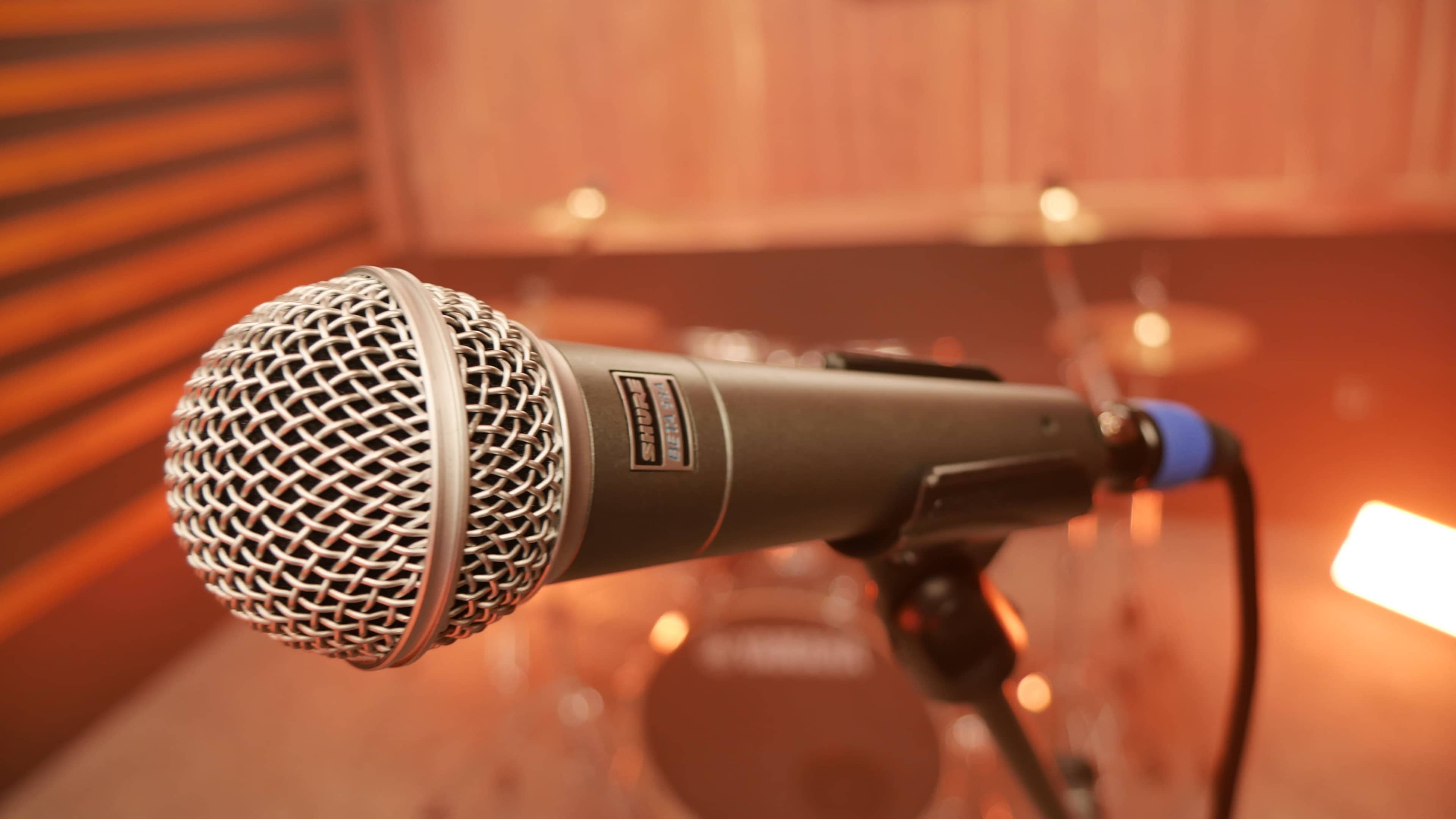 A close-up of a microphone on a stand, with blurry drum set equipment in the background.