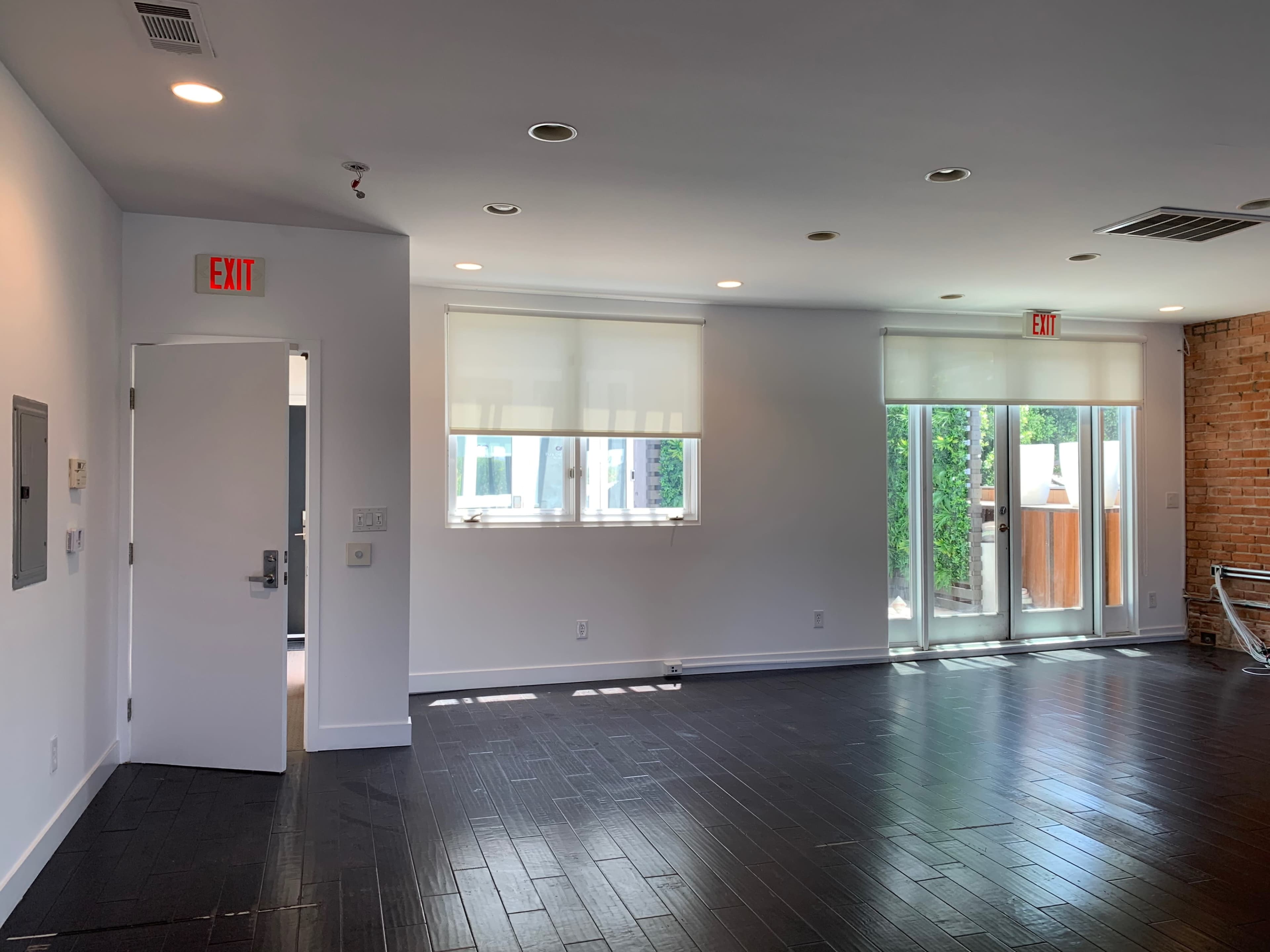 An interior space with dark wooden floors, white walls, large windows, and an exit door leading outdoors.