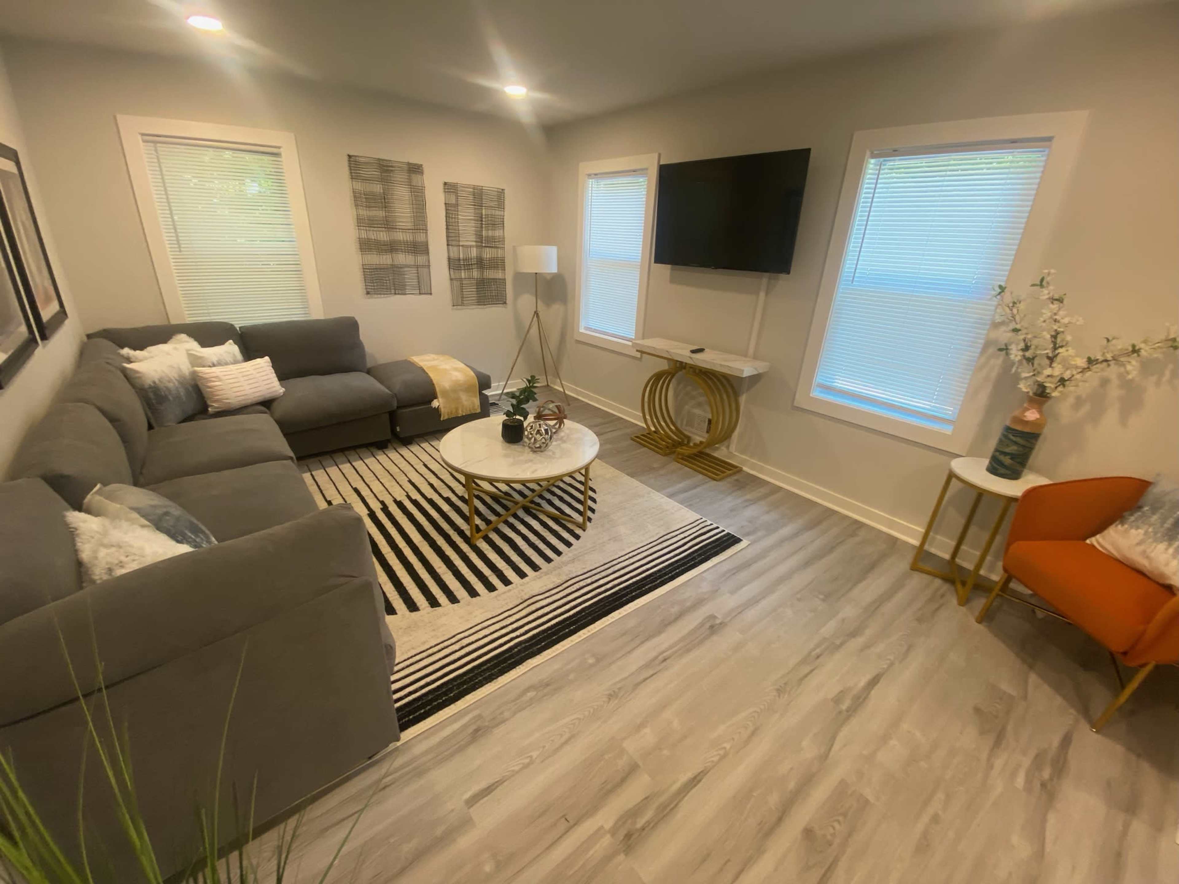 A modern living room features a gray sectional sofa, a round coffee table, a striped area rug, and a television mounted on the wall under large windows.