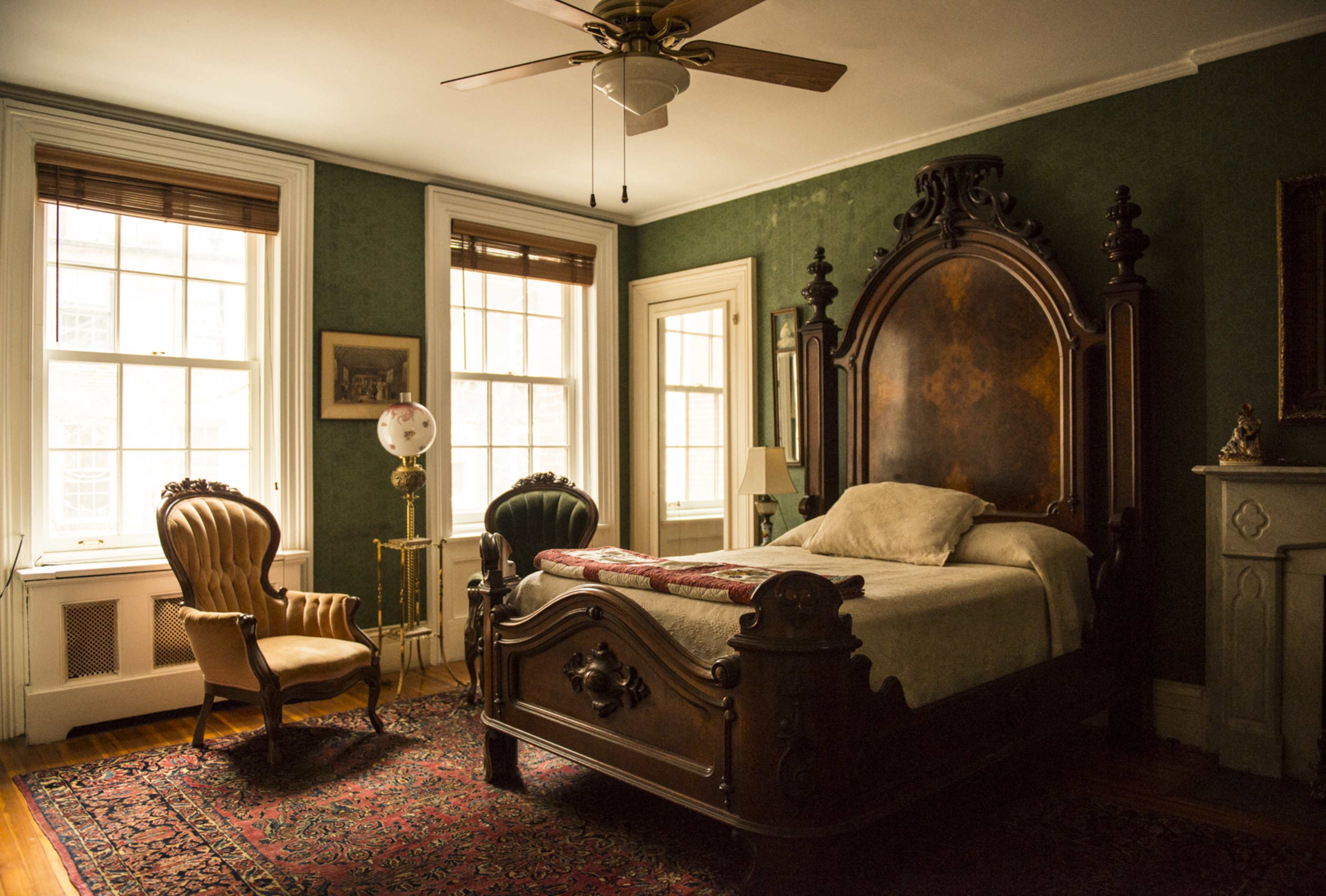 A vintage bedroom featuring a large wooden bed, a patterned rug, and an armchair near a window with wooden blinds.