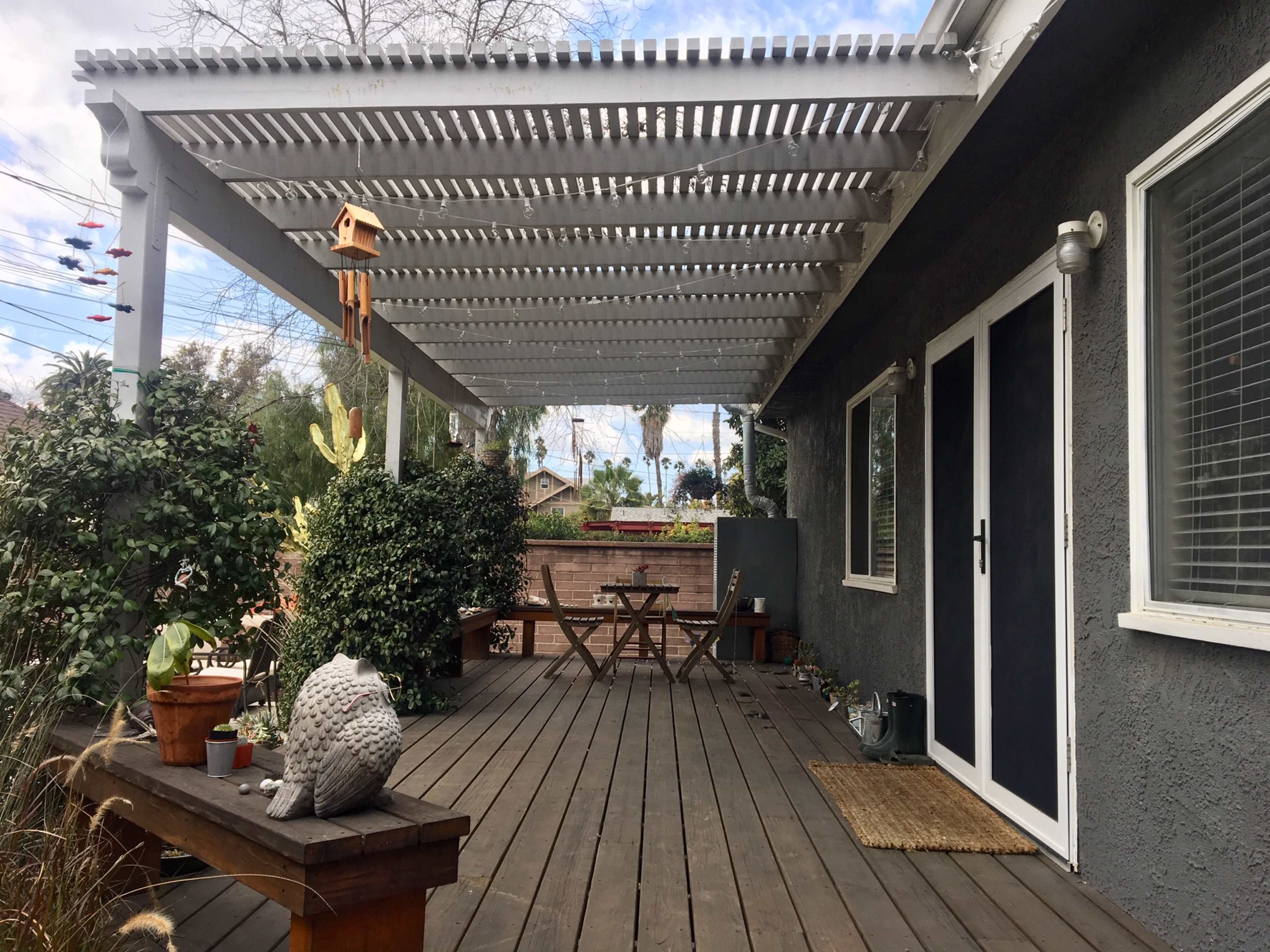 A wooden deck with a pergola overhead features potted plants, a picnic table, and a decorative owl statue.